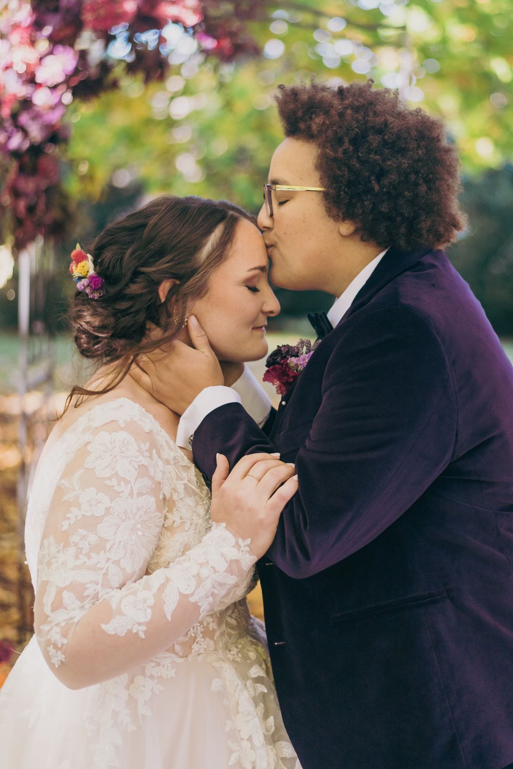 Spouse in purple velvet suit kissing bride in lace gown on the cheek at outdoor ceremony.