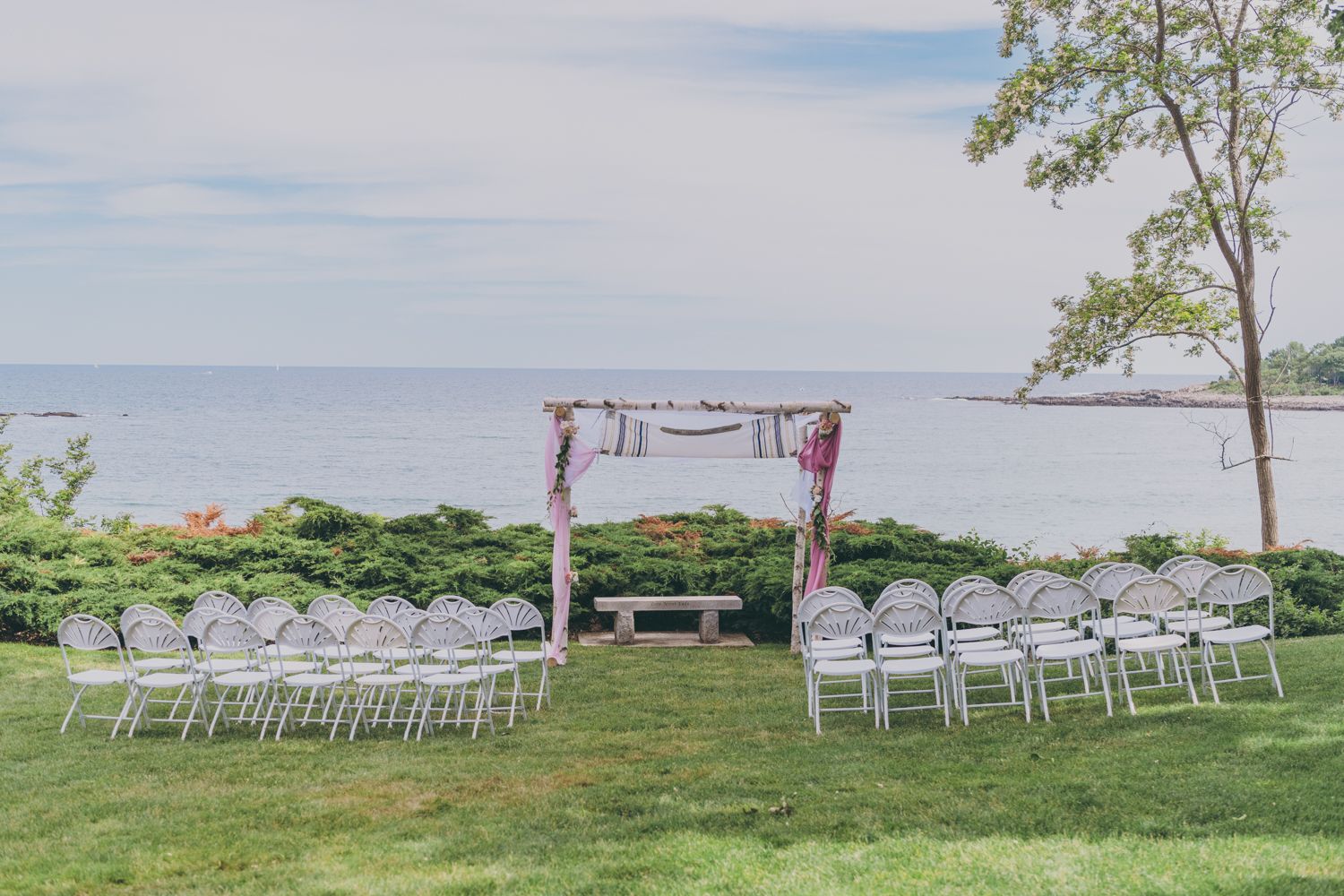 Elegant outdoor wedding ceremony setup with white chairs and floral arch overlooking a serene ocean coastline.