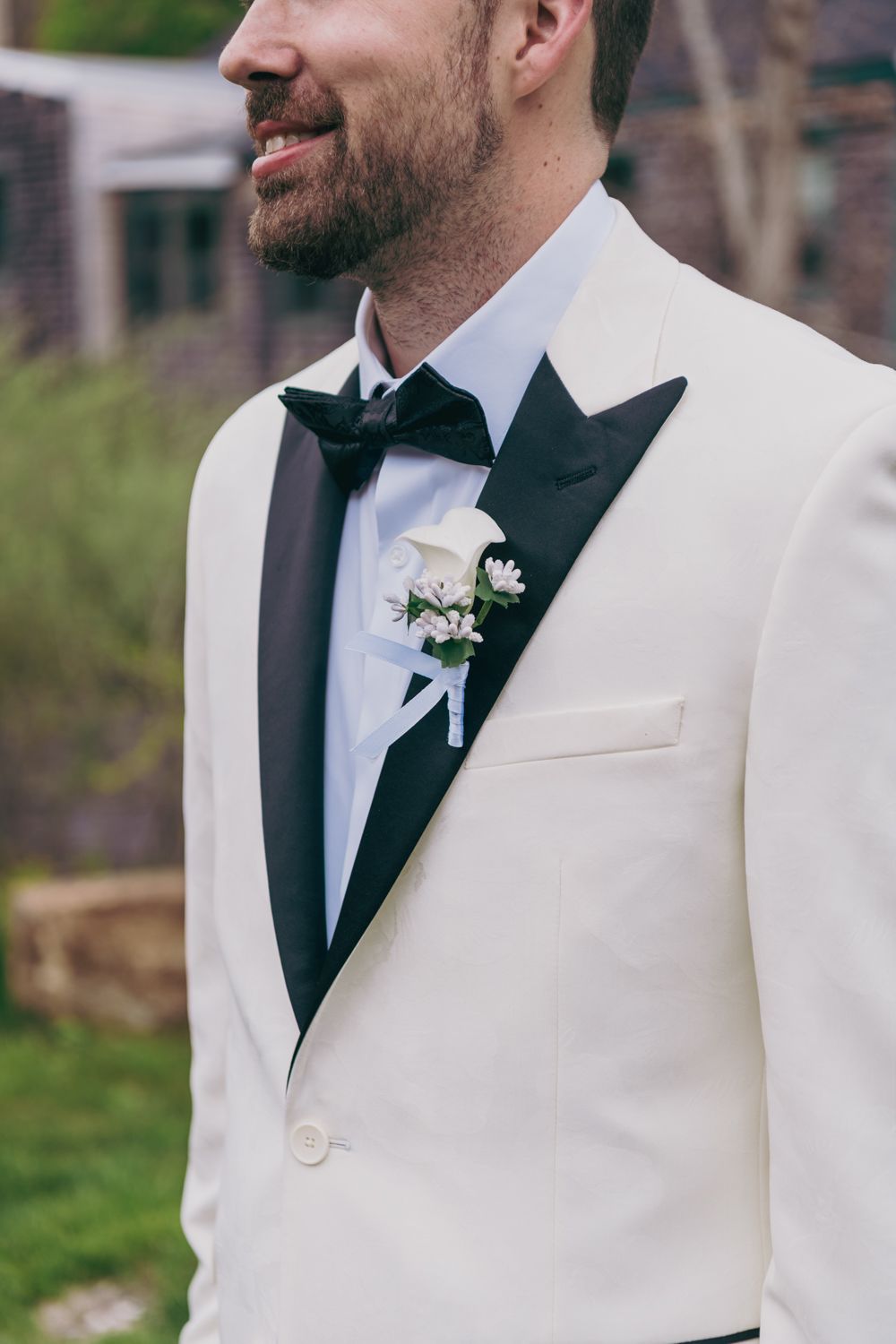 Groom in white tuxedo with black lapels, bow tie, and white floral boutonniere smiling outdoors.