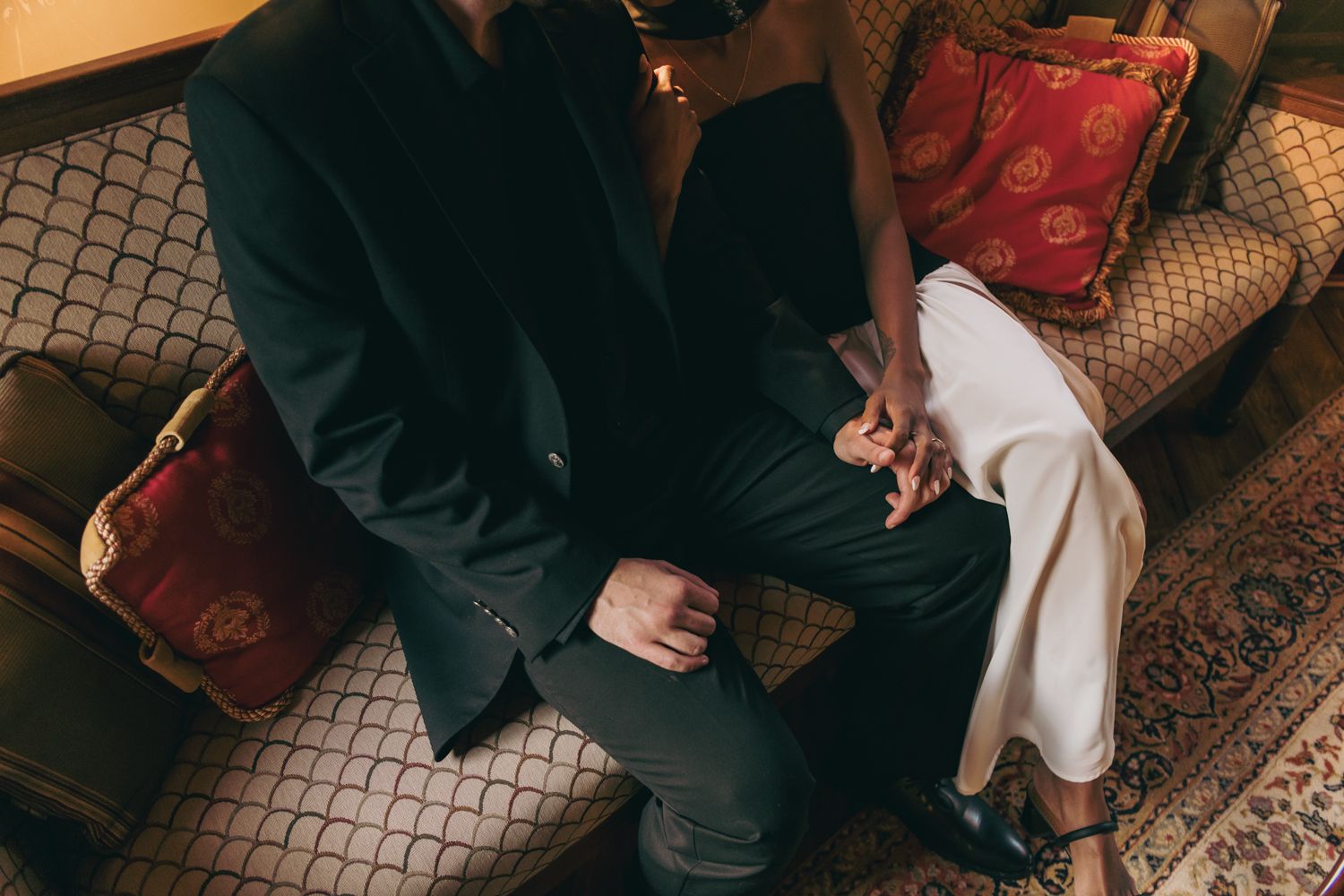Couple sitting closely on an ornate sofa, holding hands, dressed in elegant black and white attire.