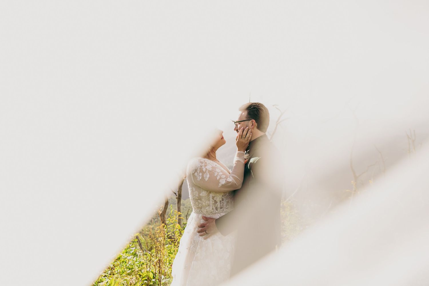 Bride in lace wedding dress leaning against a white wall with greenery visible below.