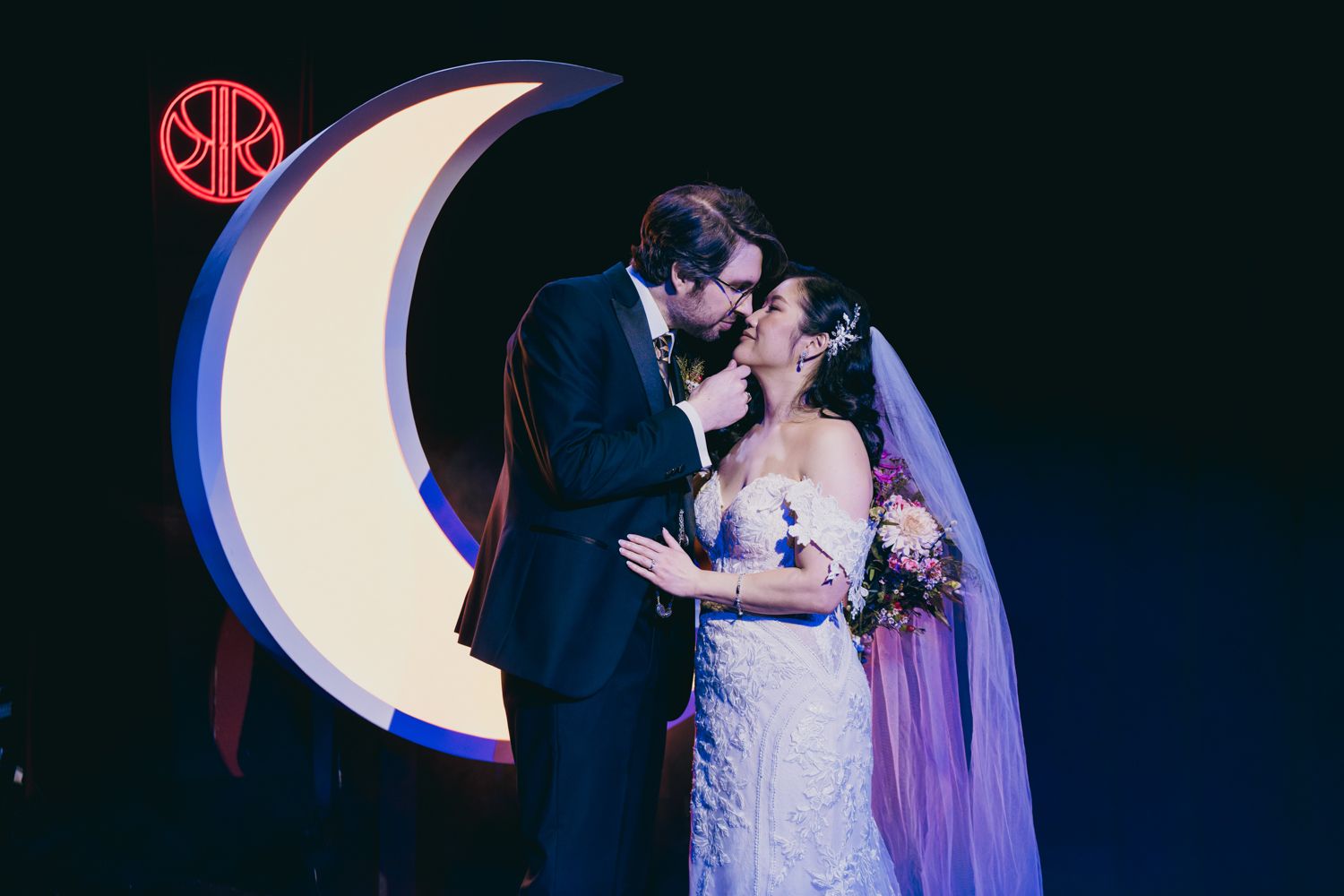Bride and groom share a kiss in front of an illuminated crescent moon wedding backdrop.