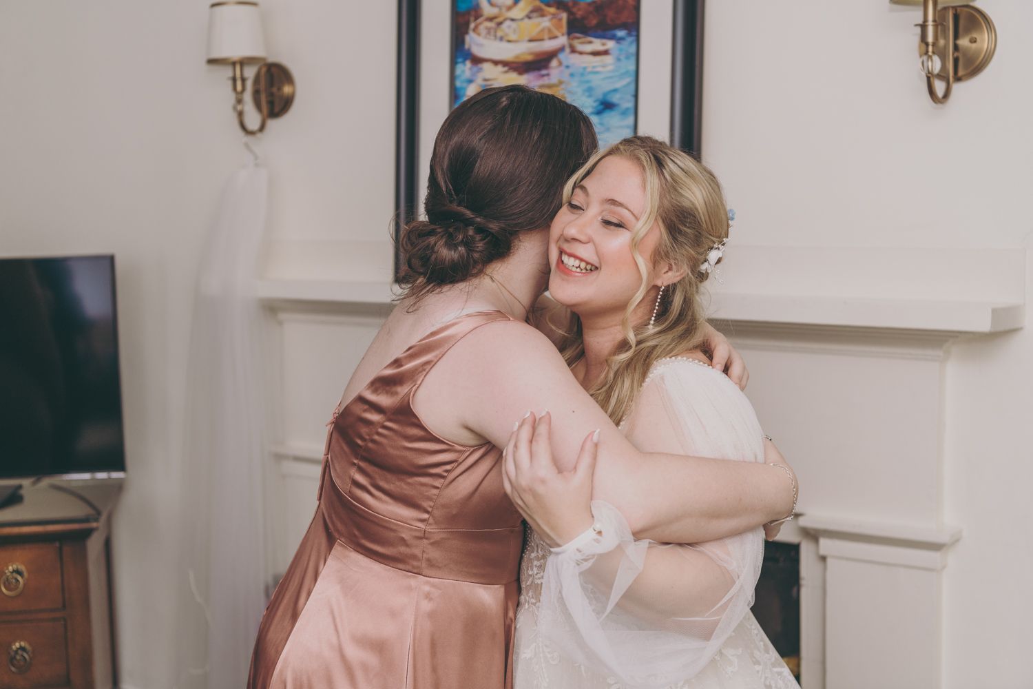 A bride in a white gown shares a warm hug with a bridesmaid in a pink satin dress during wedding preparations.