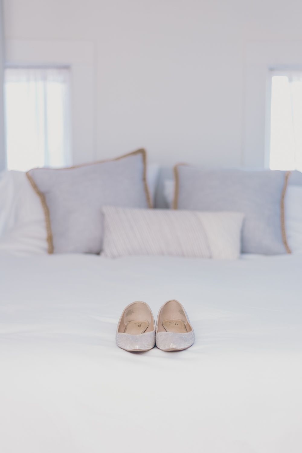 Elegant white bridal flats displayed on a neatly made bed with soft gray pillows.