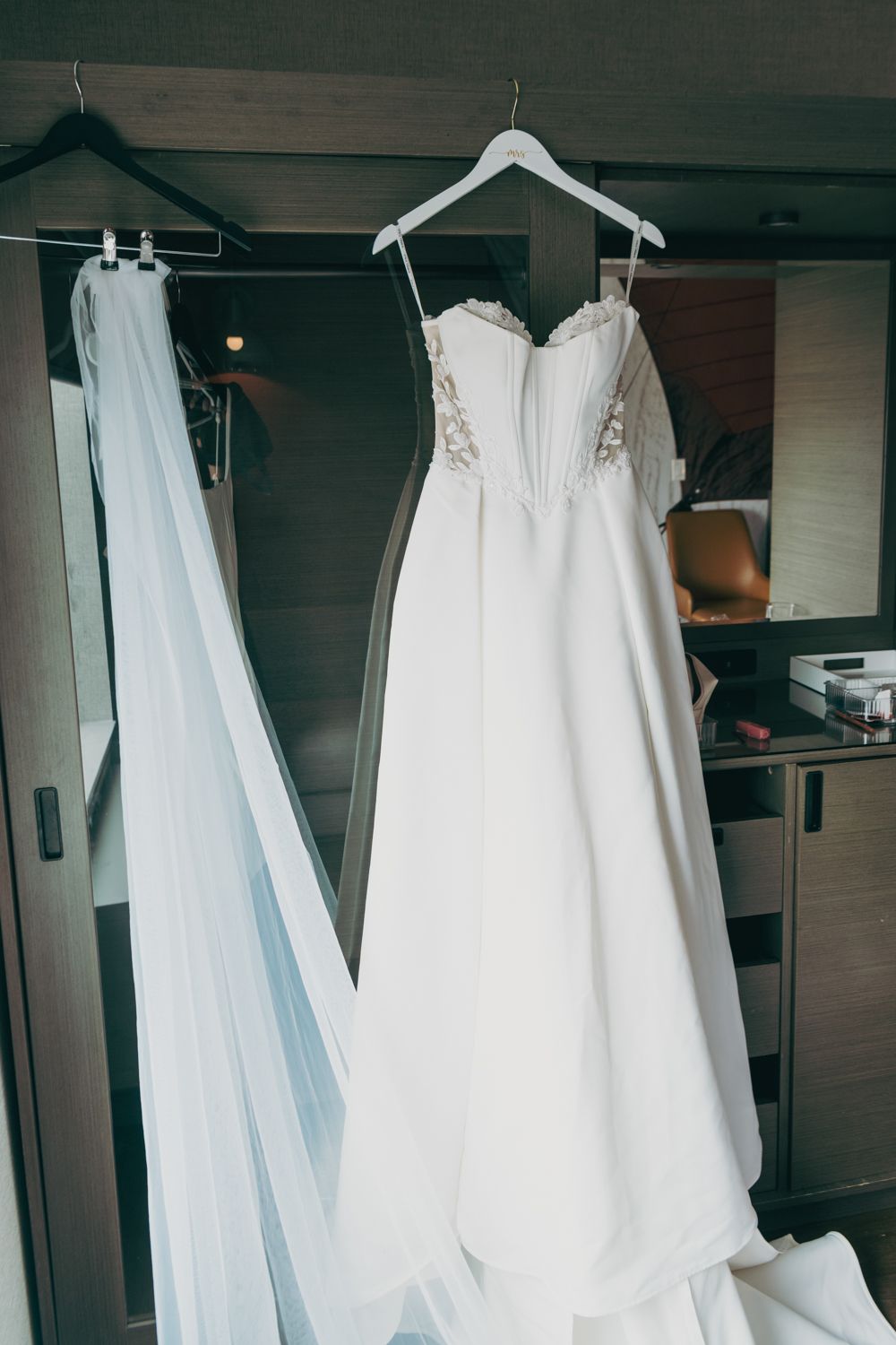 Elegant white A-line wedding dress with lace bodice hanging on a white hanger in a hotel room.
