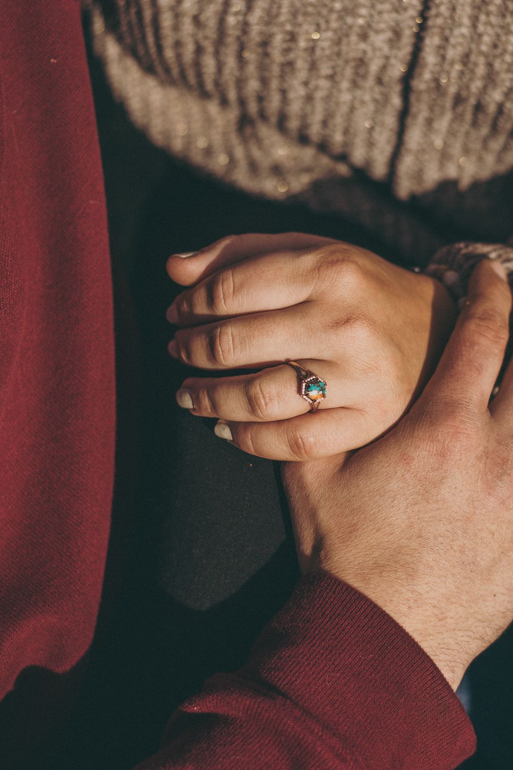 Close-up of two hands holding, woman wearing a gold ring with a teal gemstone, wearing cozy knit sweaters.