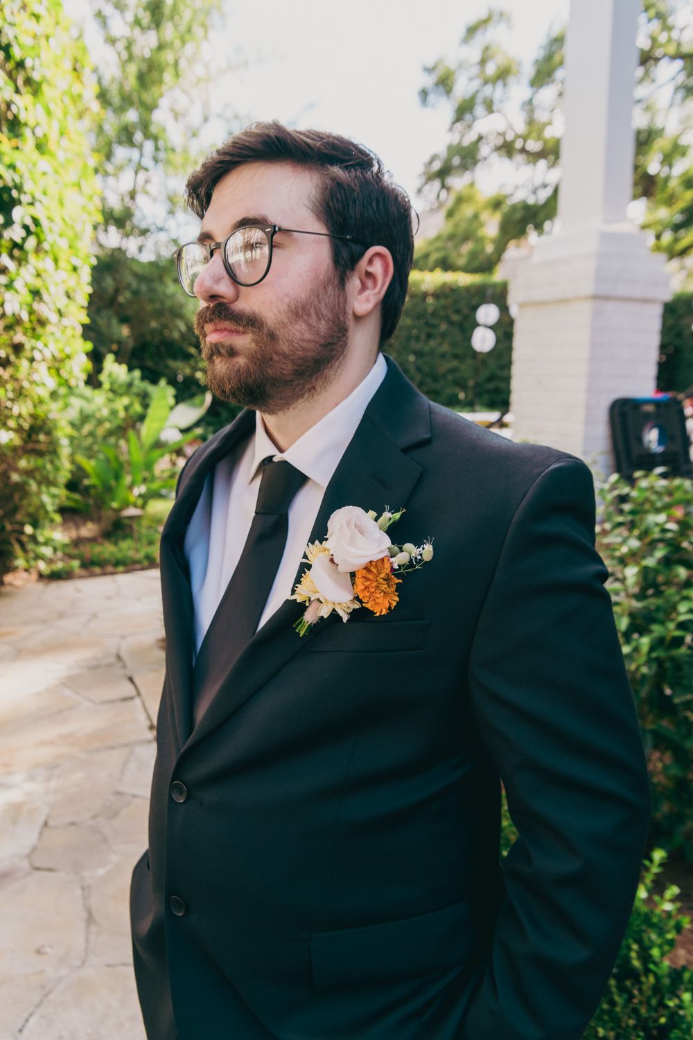 Dapper groom in dark suit with floral boutonniere of roses and orange flowers poses in a garden setting.