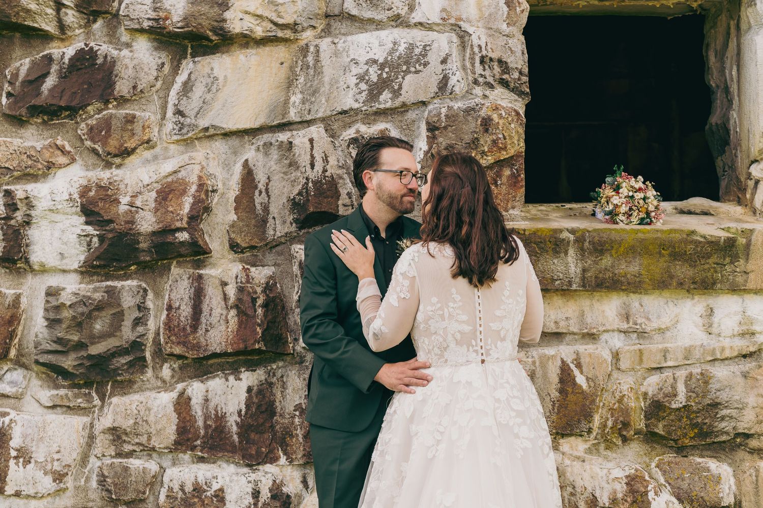 Bride and groom share a tender kiss against a rustic stone wall, with a floral bouquet resting on the ledge.