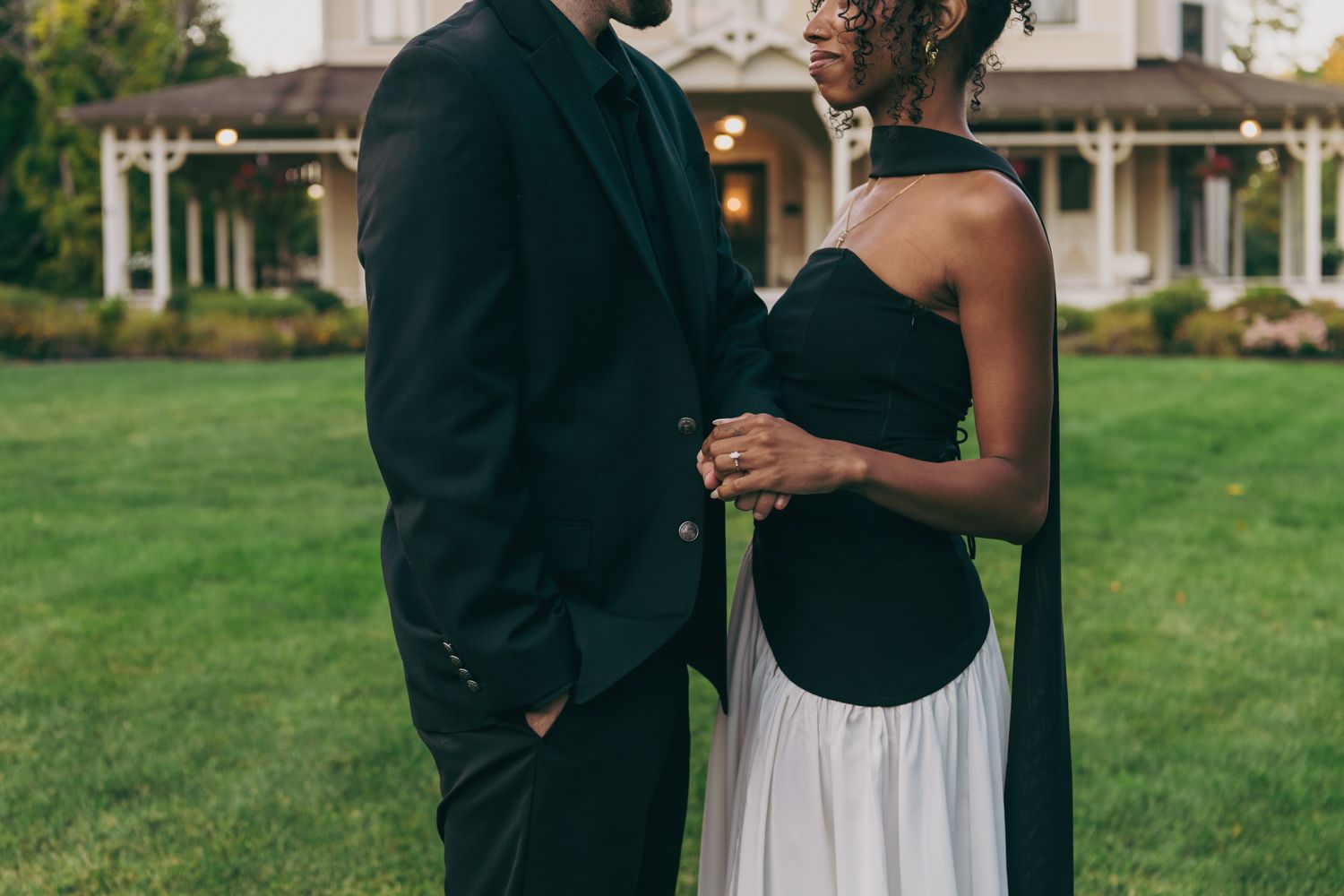 Interracial couple standing close together on lawn wearing elegant black and white formal attire.