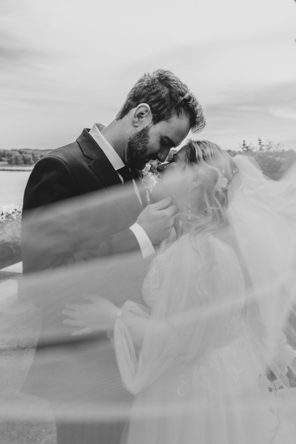 Black and white photo of bride and groom sharing a romantic kiss under flowing wedding veil.