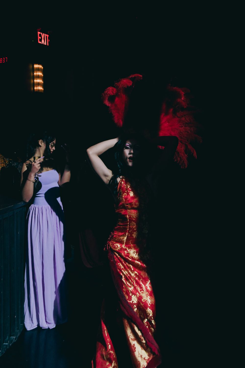Woman in red Chinese qipao dress with feathered headpiece poses in dark venue near exit sign.