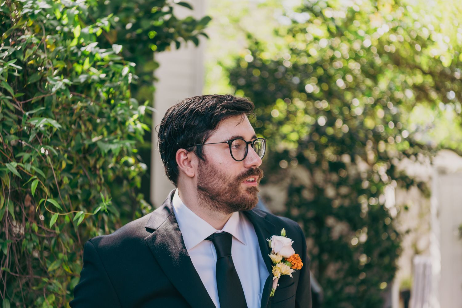 Groom in a black suit with boutonniere stands outdoors among green foliage on his wedding day.