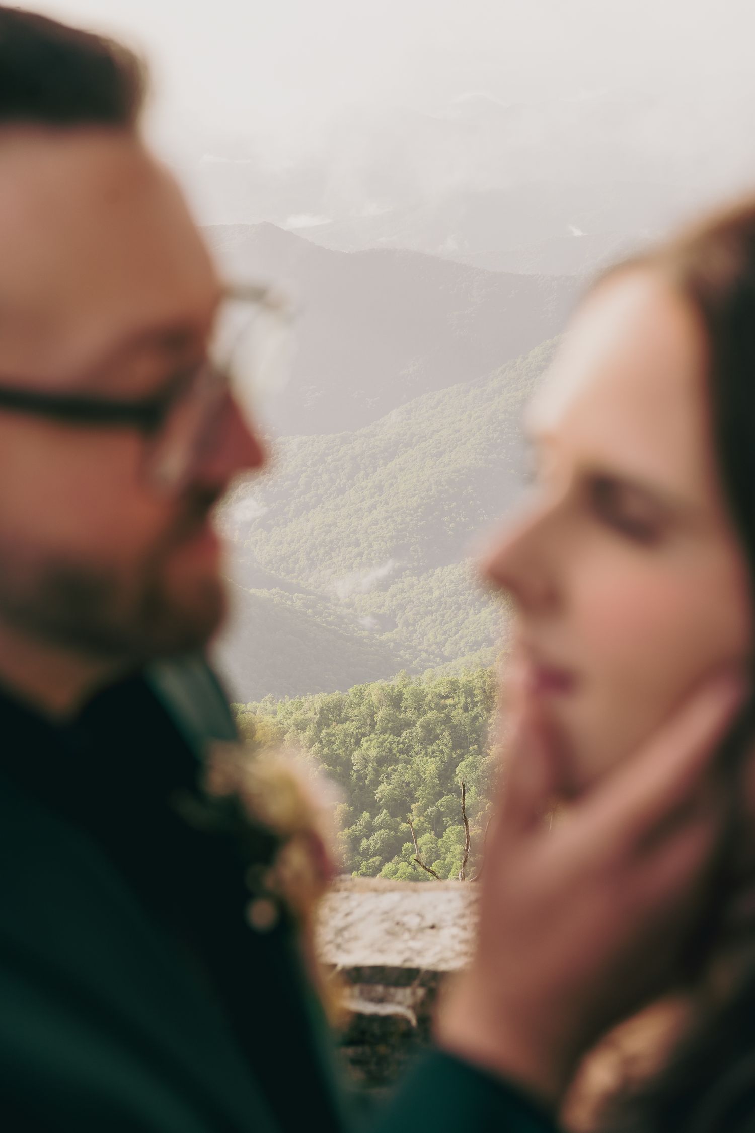 A couple shares an intimate moment with a lush mountain forest backdrop, the woman's face gently cupped in soft focus.
