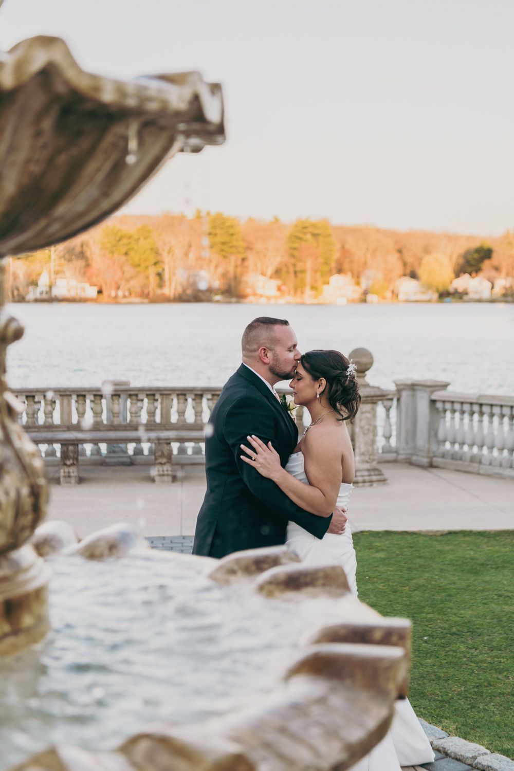A bride and groom share a romantic embrace on an elegant stone terrace overlooking a serene lake at golden hour.