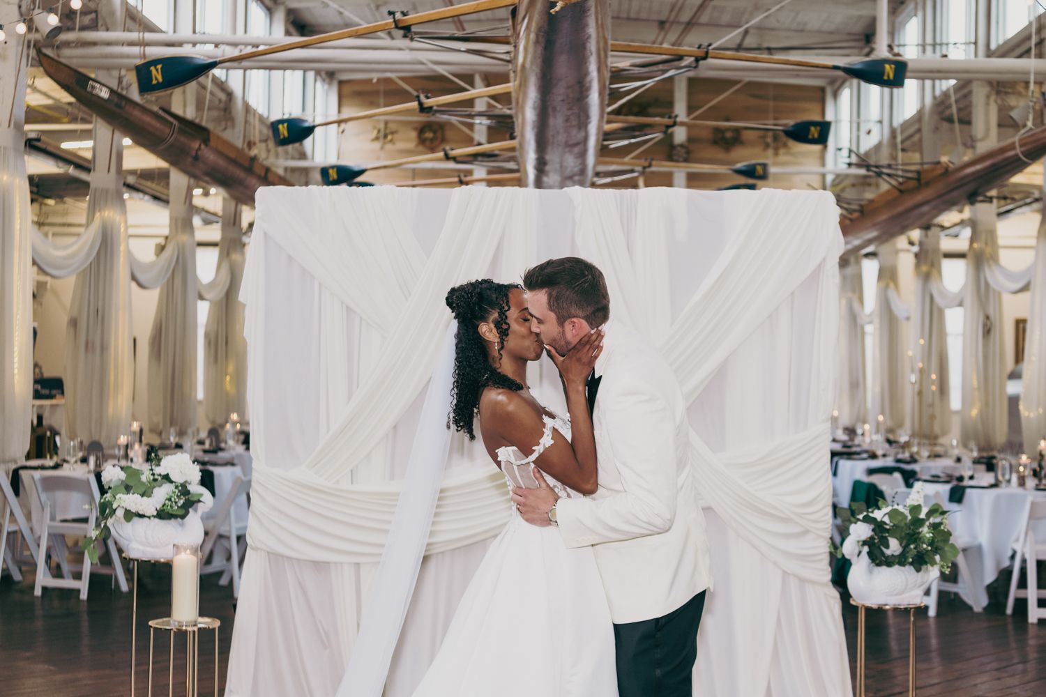 Newlywed interracial couple shares first kiss under white draped arch in elegant industrial wedding venue.