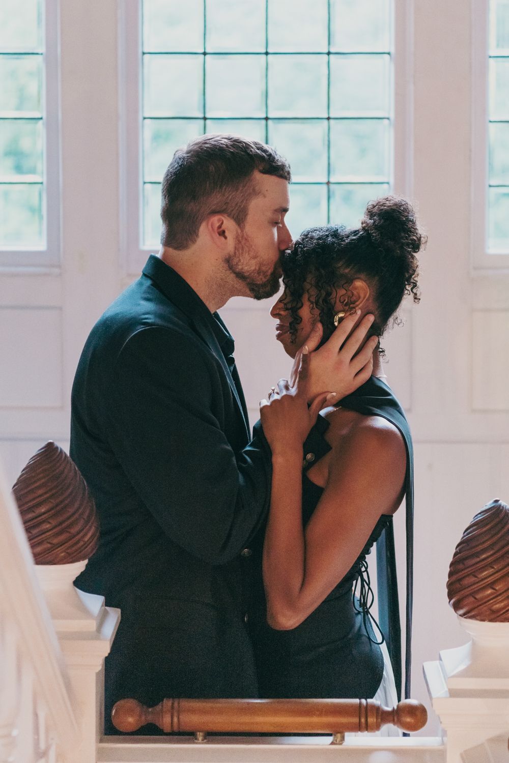Romantic interracial couple sharing a tender kiss in a bright white room with large windows.