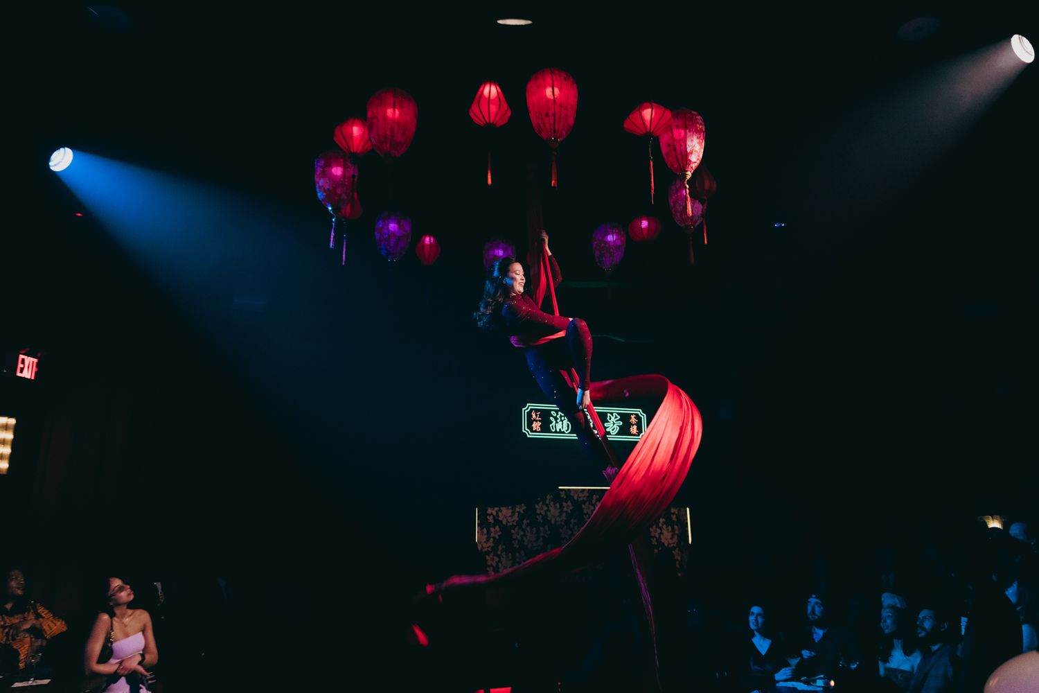 Aerial silk performer suspended above crowd under red lanterns at wedding reception.