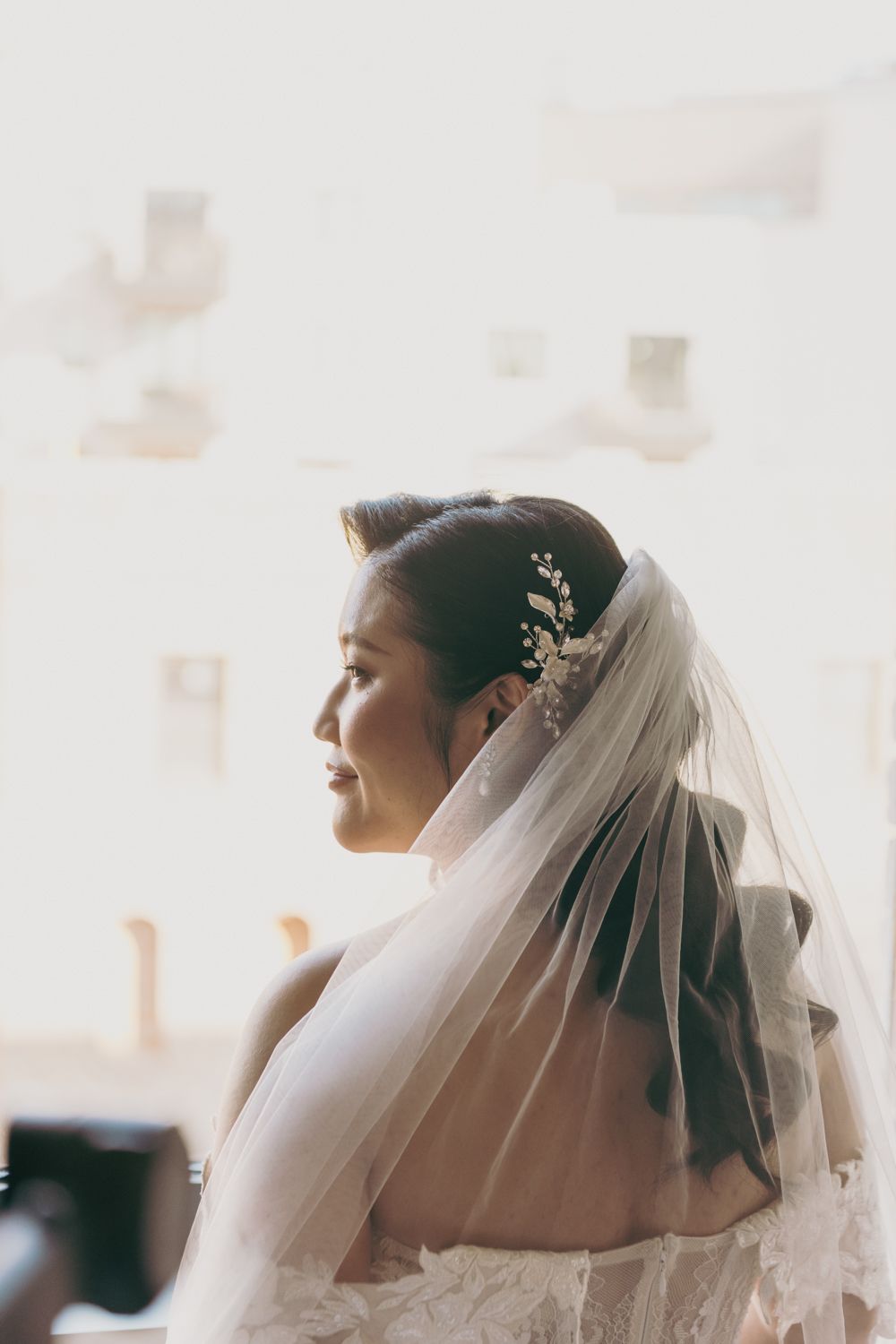 A bride in a white gown and veil with a floral hair accessory gazes softly to the side in elegant profile.