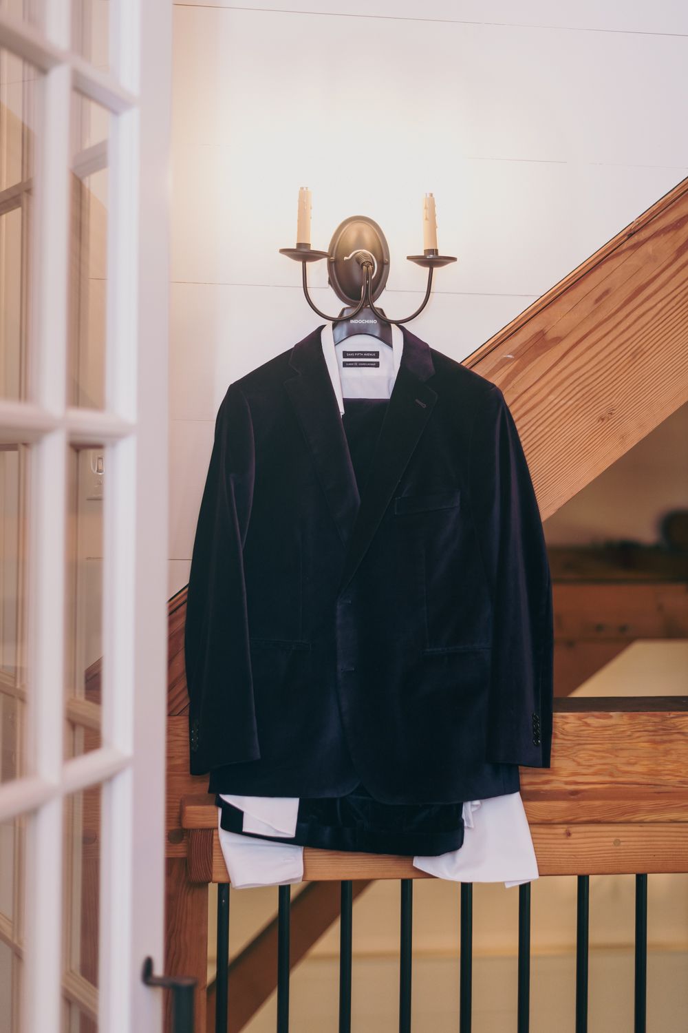 Dark navy velvet groom suit jacket hanging on vintage brass candelabra wall sconce on wooden staircase.