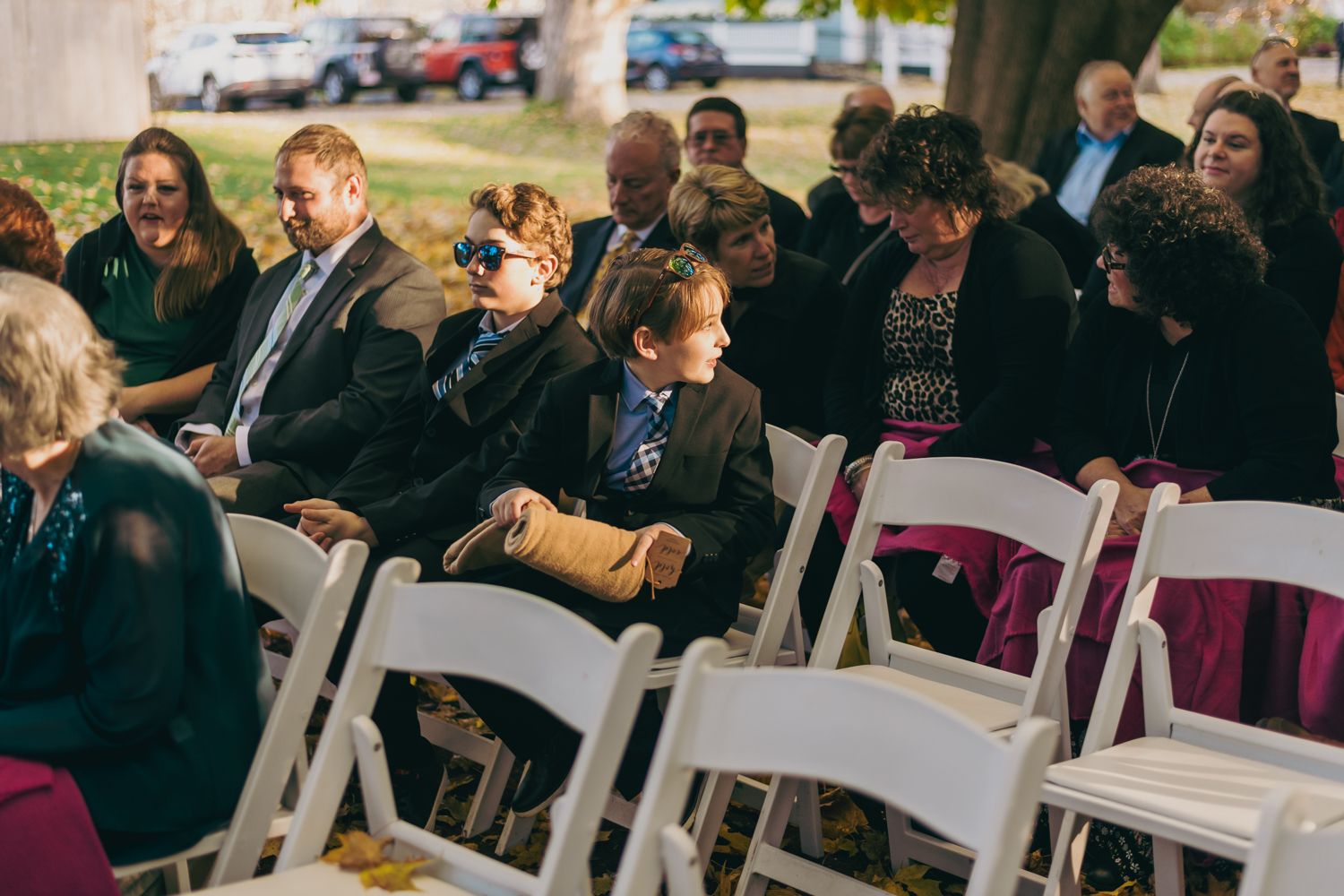Wedding guests seated in white chairs at outdoor ceremony on sunny autumn day.