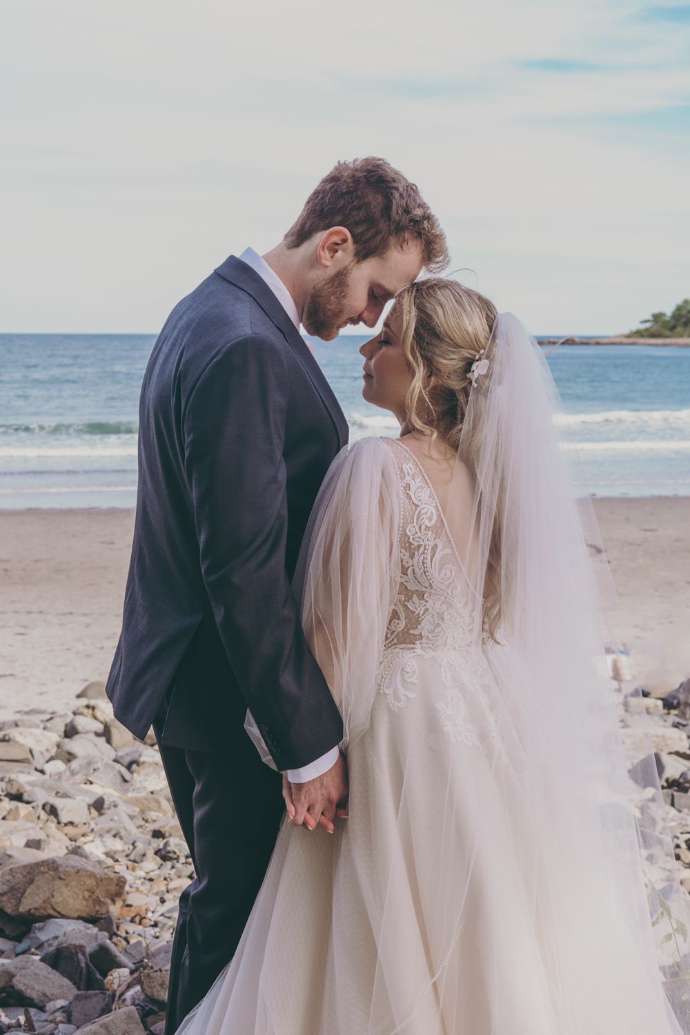 A bride and groom touch foreheads tenderly while holding hands on a rocky beach after their wedding.