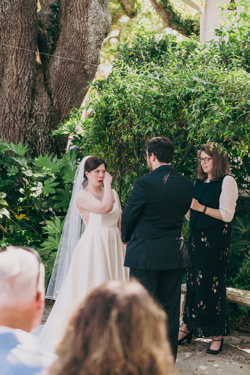 Emotional bride wipes tears during outdoor wedding ceremony as officiant and groom look on.