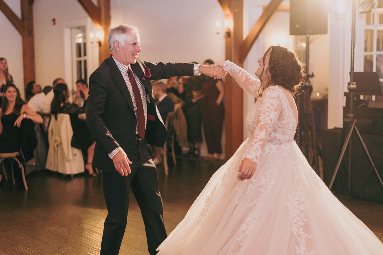 Bride in lace gown dancing with older man at elegant indoor wedding reception.