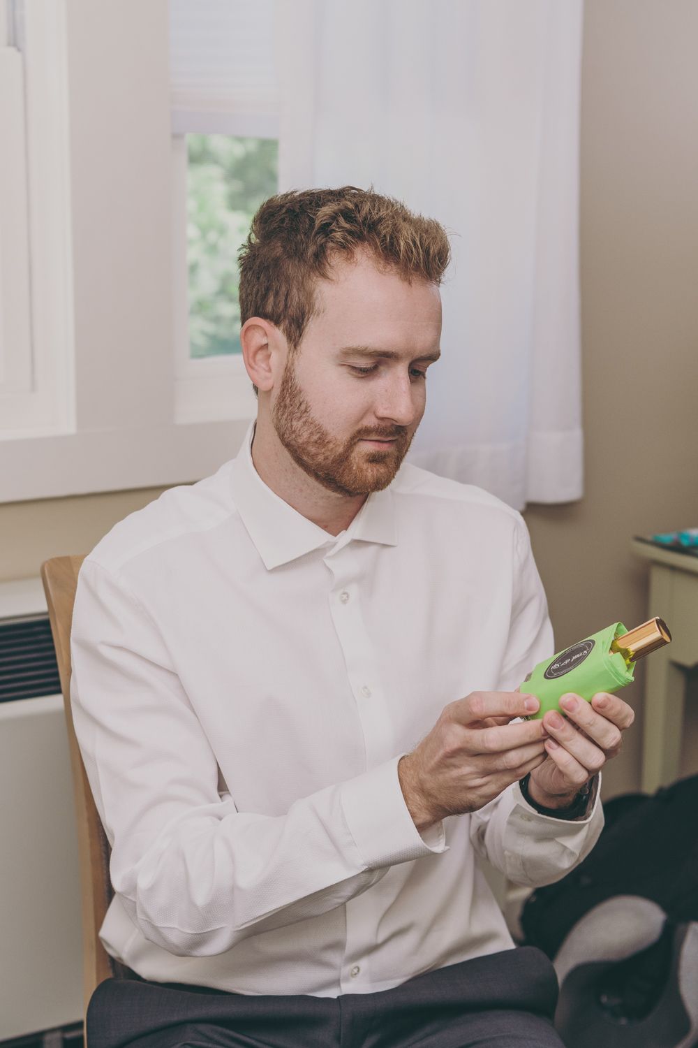 Groom in white dress shirt examining green cologne bottle while seated before his wedding ceremony.