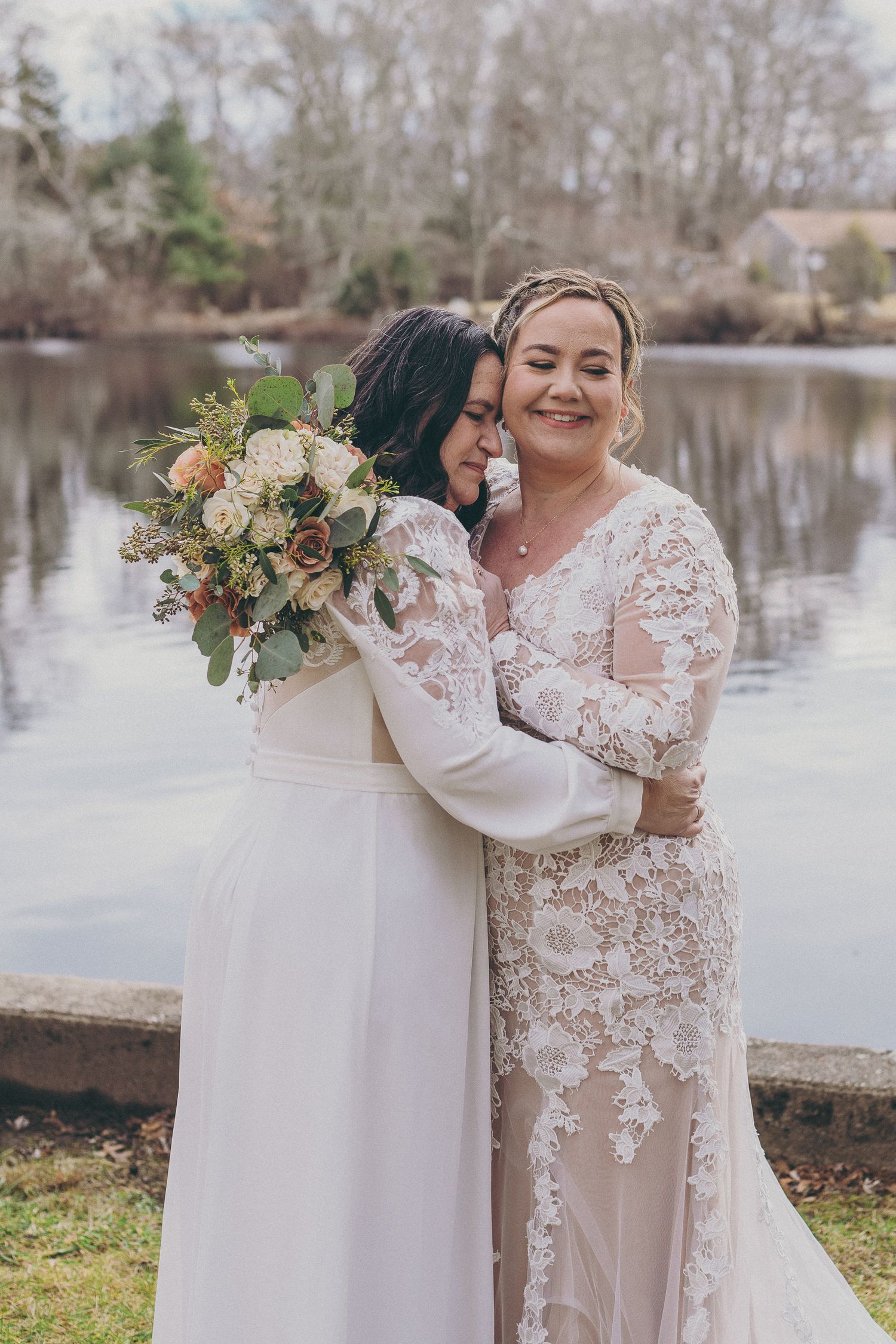 Two brides in elegant lace wedding gowns embrace warmly while holding a floral bouquet near a serene lake backdrop.
