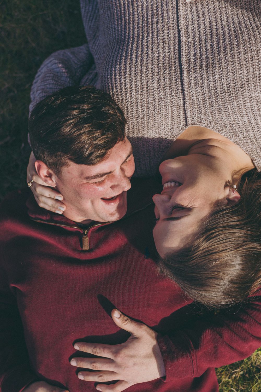A happy couple lying on grass, laughing and embracing, wearing cozy red and gray sweaters in an outdoor setting.
