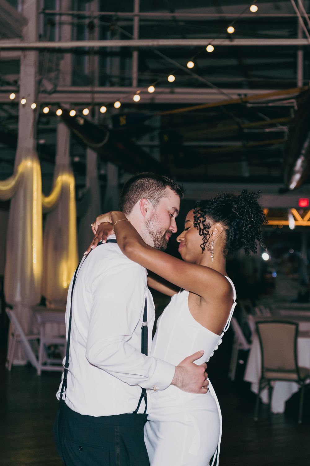 Newlyweds sharing intimate slow dance embrace under string lights at indoor wedding reception venue.