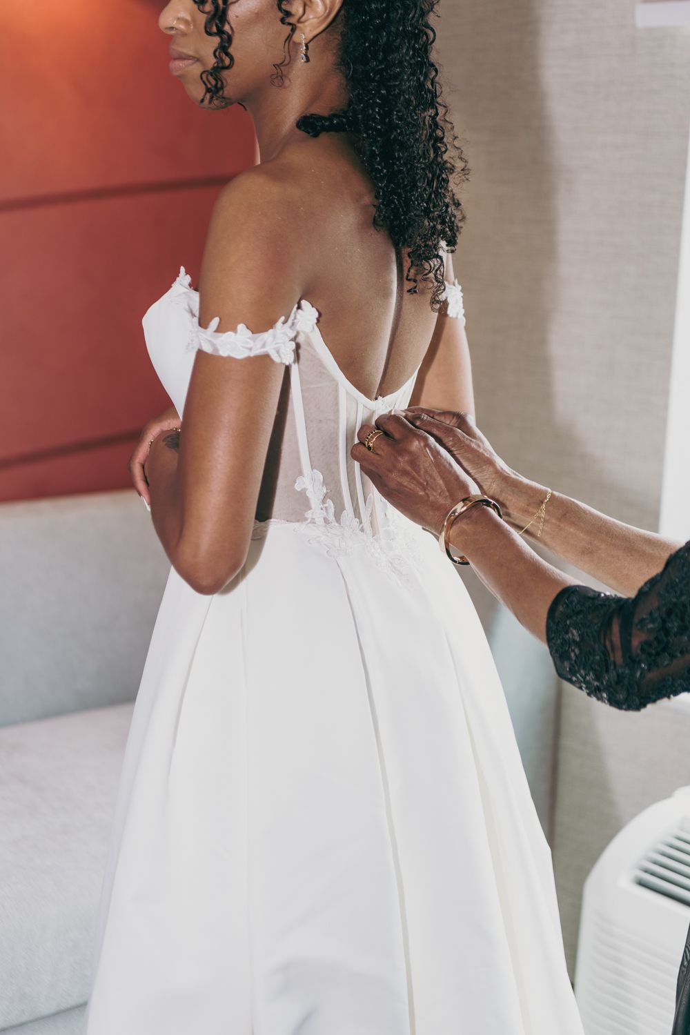 A bride in a white off-shoulder wedding gown being helped with the back of her dress by an assistant.