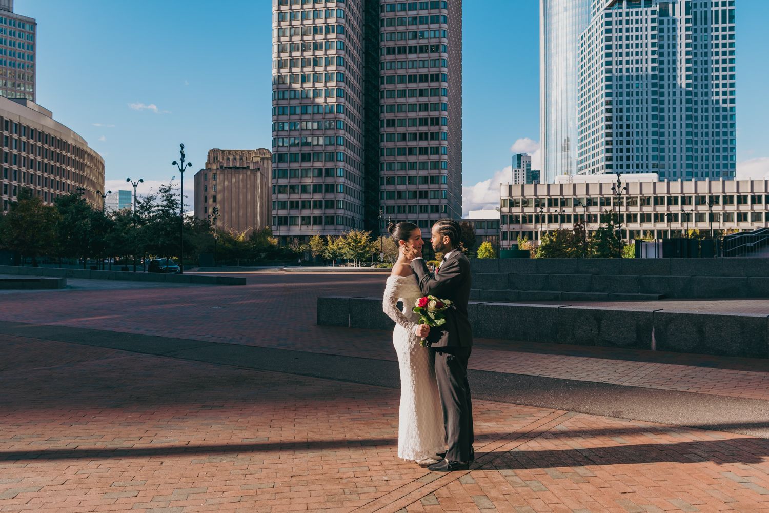 Bride and groom embrace in front of city skyscrapers on a sunny day in an urban plaza with brick pavement.