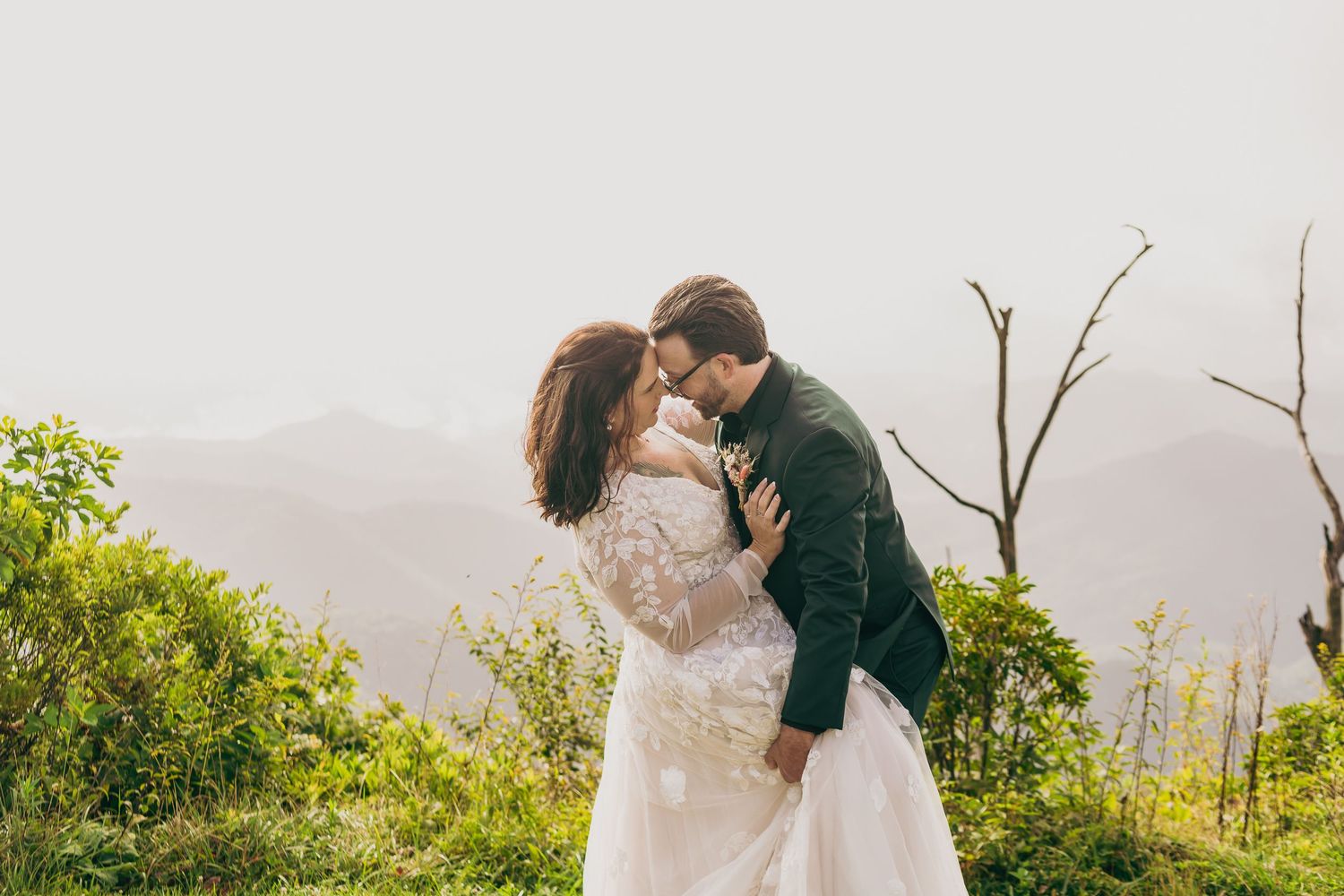 Bride and groom sharing a romantic kiss on a misty mountain overlook surrounded by lush greenery in wedding attire.