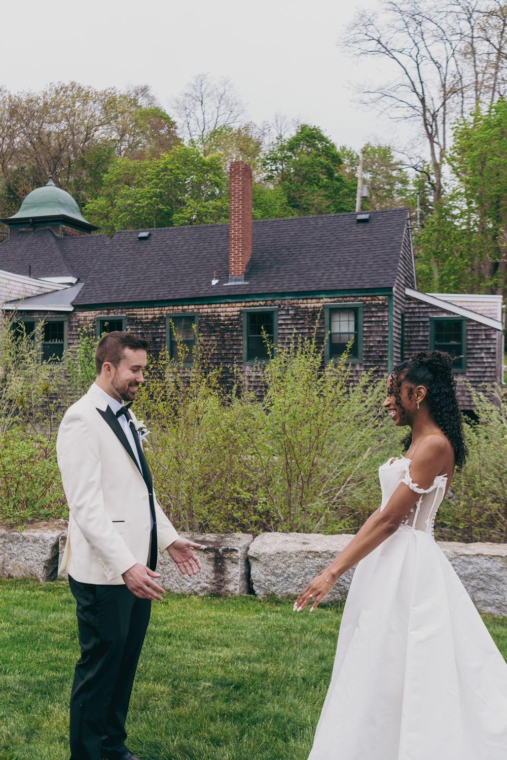 Interracial couple sharing joyful first look moment in garden, bride in white gown, groom in white tuxedo.