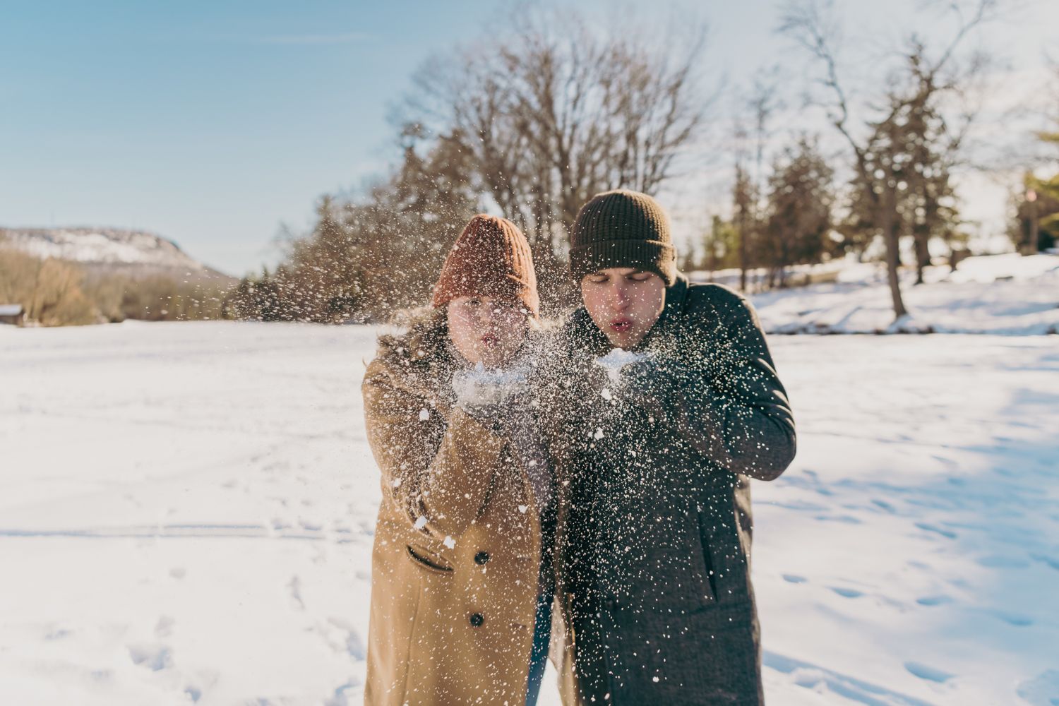 Couple blowing snow from hands in snowy winter landscape, wearing coats and hats on sunny day.