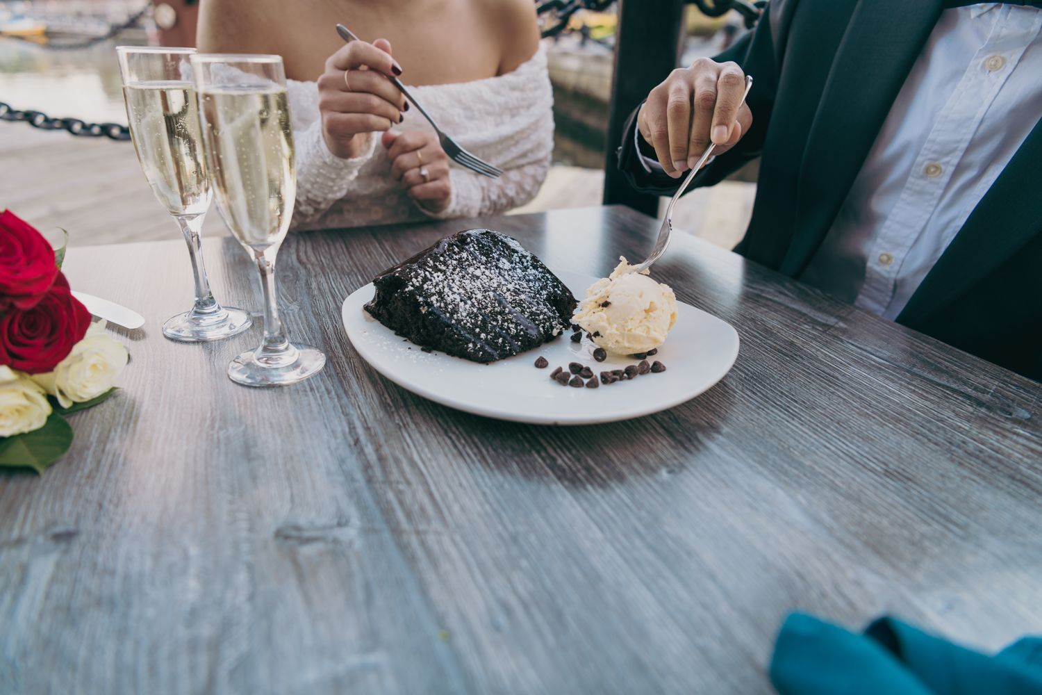 Newlyweds share a chocolate dessert with champagne flutes and red roses on a waterfront restaurant table.