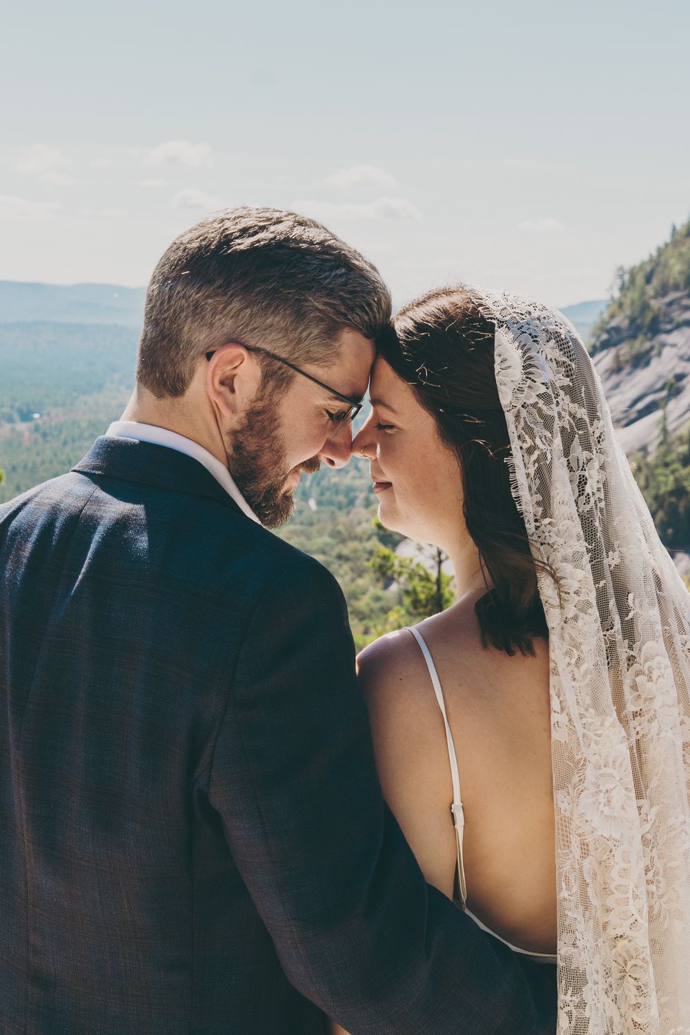 A bride and groom touch foreheads tenderly outdoors, with a lace veil and mountain scenery in the background.