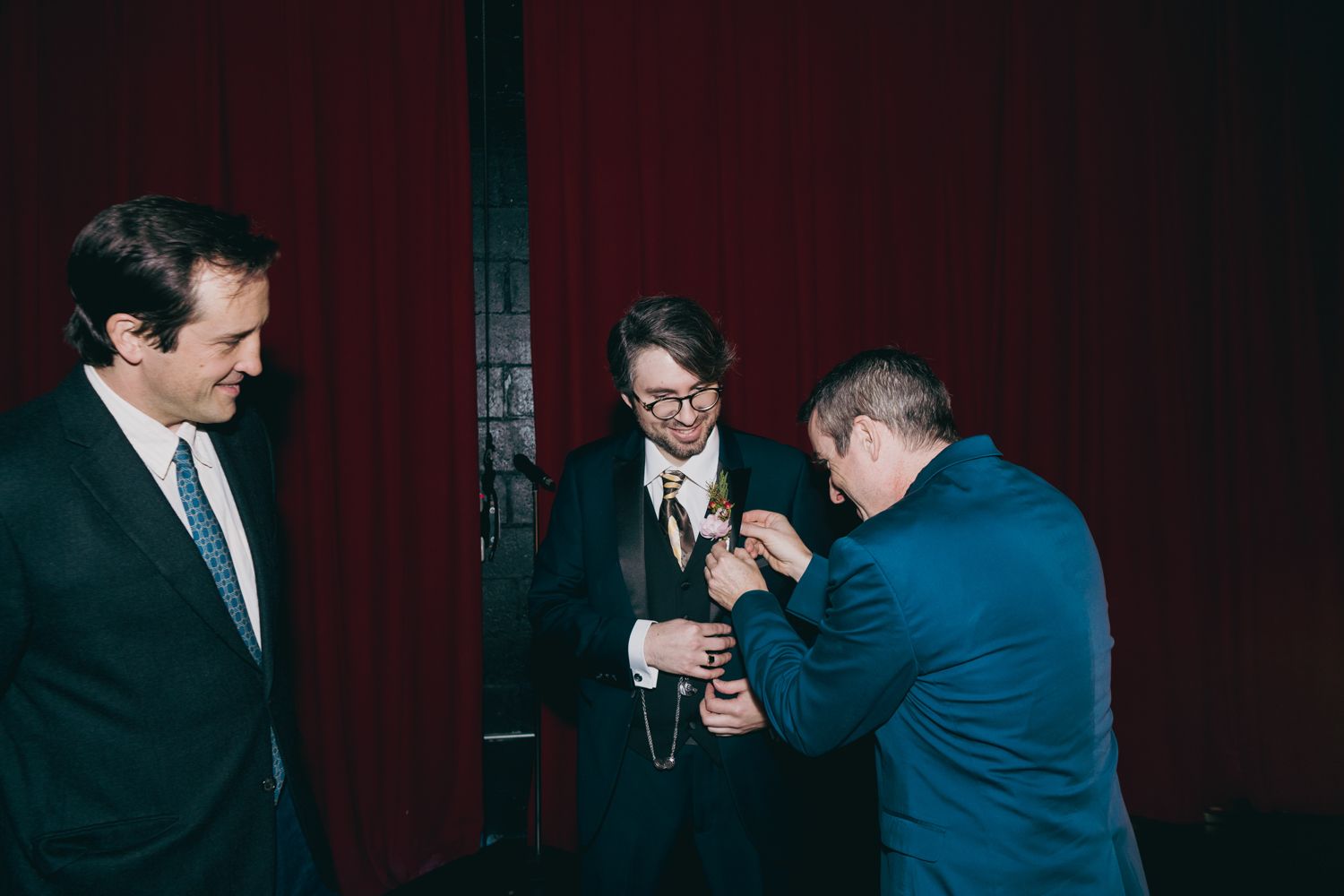 Three men in suits share a moment as one pins a boutonniere on another at a formal wedding reception.