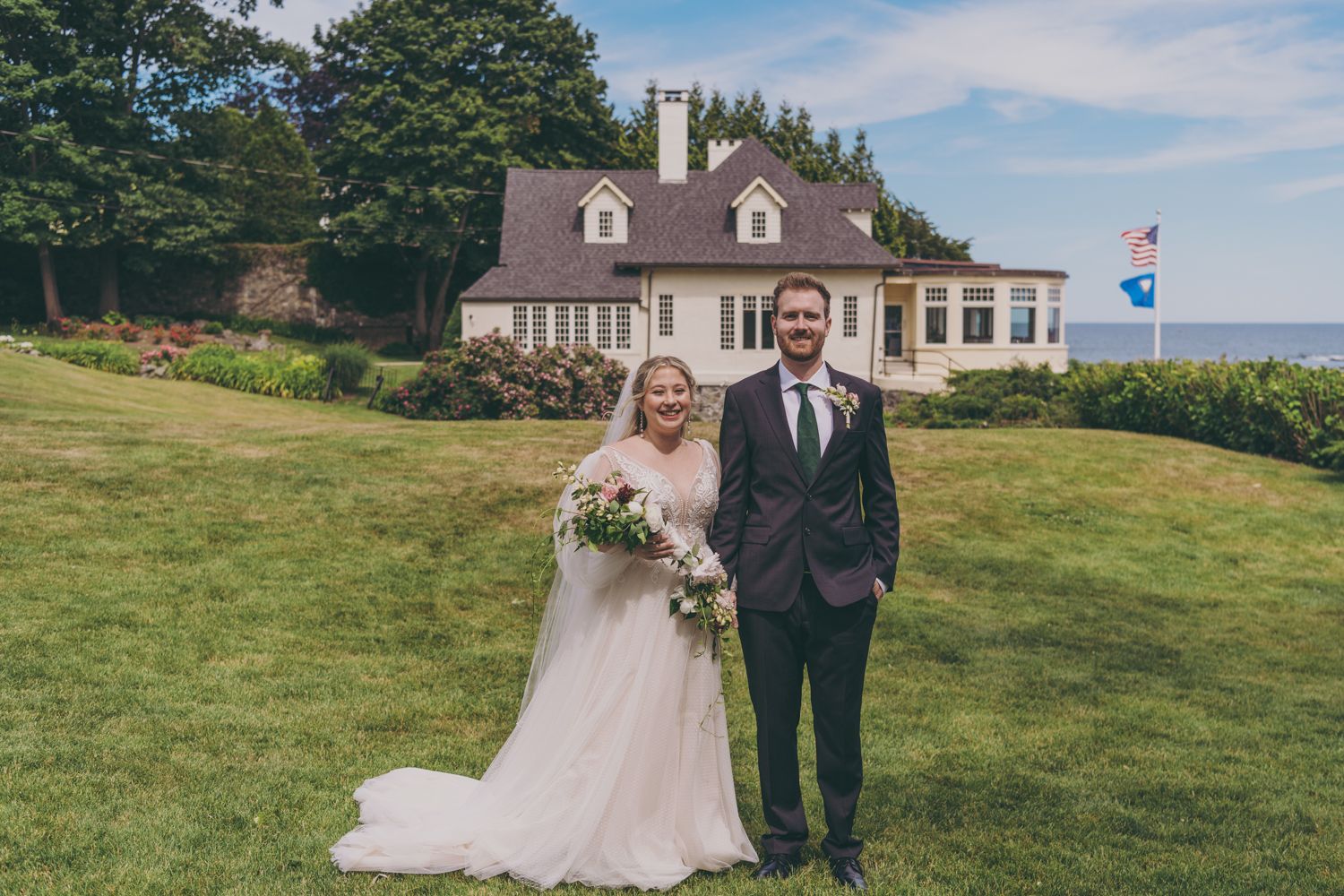 Bride and groom posing together on green lawn in front of elegant coastal estate home.