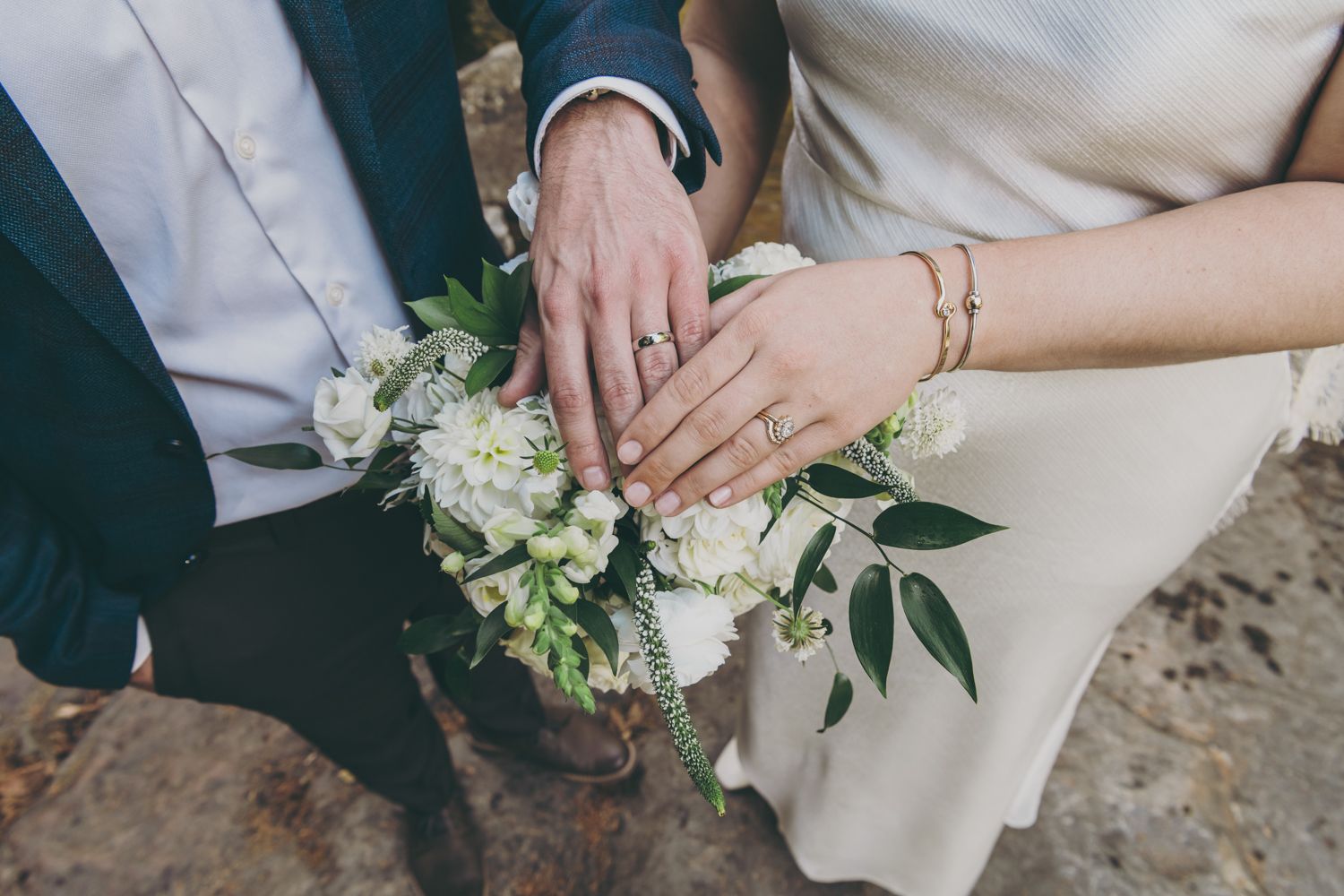 Newlyweds display wedding rings over a white floral bouquet with greenery.