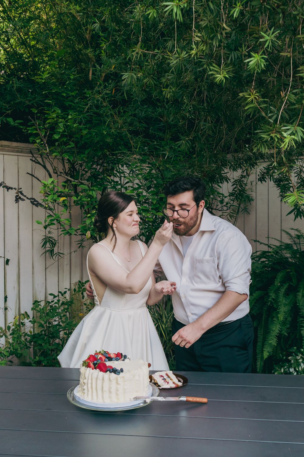 Bride feeds groom a piece of their berry-topped white wedding cake at an outdoor garden reception.