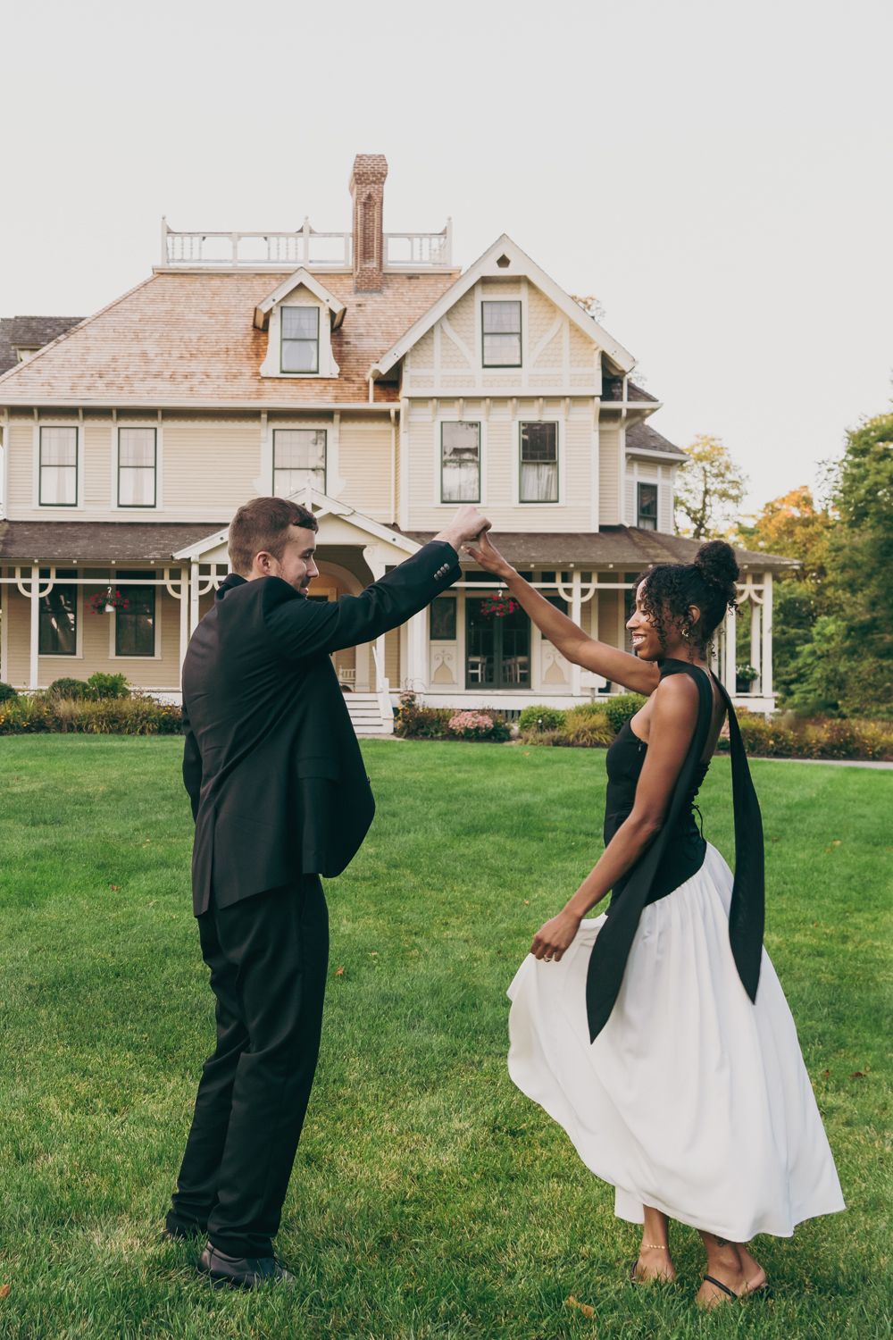 Interracial couple dancing on green lawn in front of Victorian house, man in black suit, woman in black and white dress.