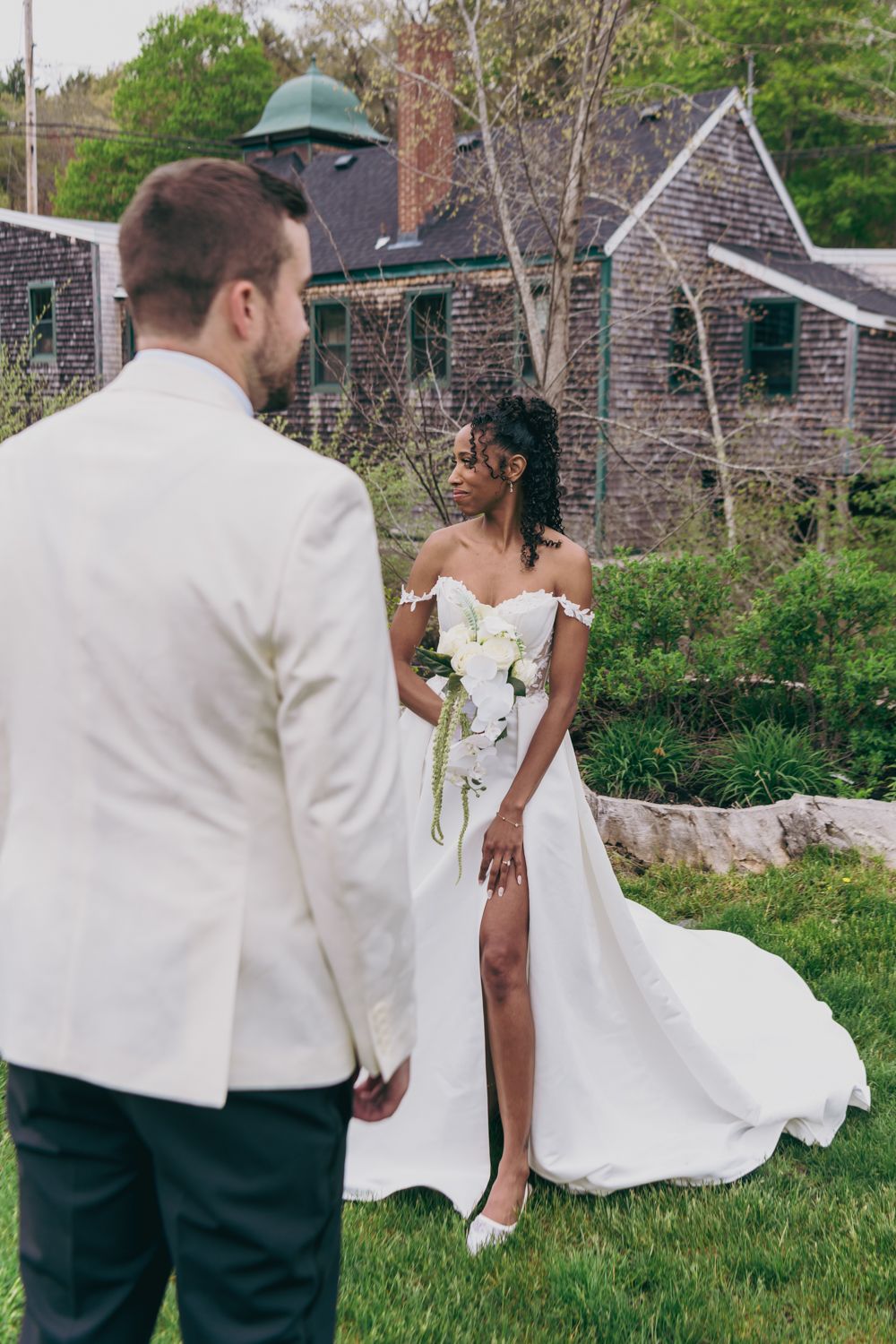 Bride in off-shoulder white gown with slit holding bouquet facing groom outdoors.