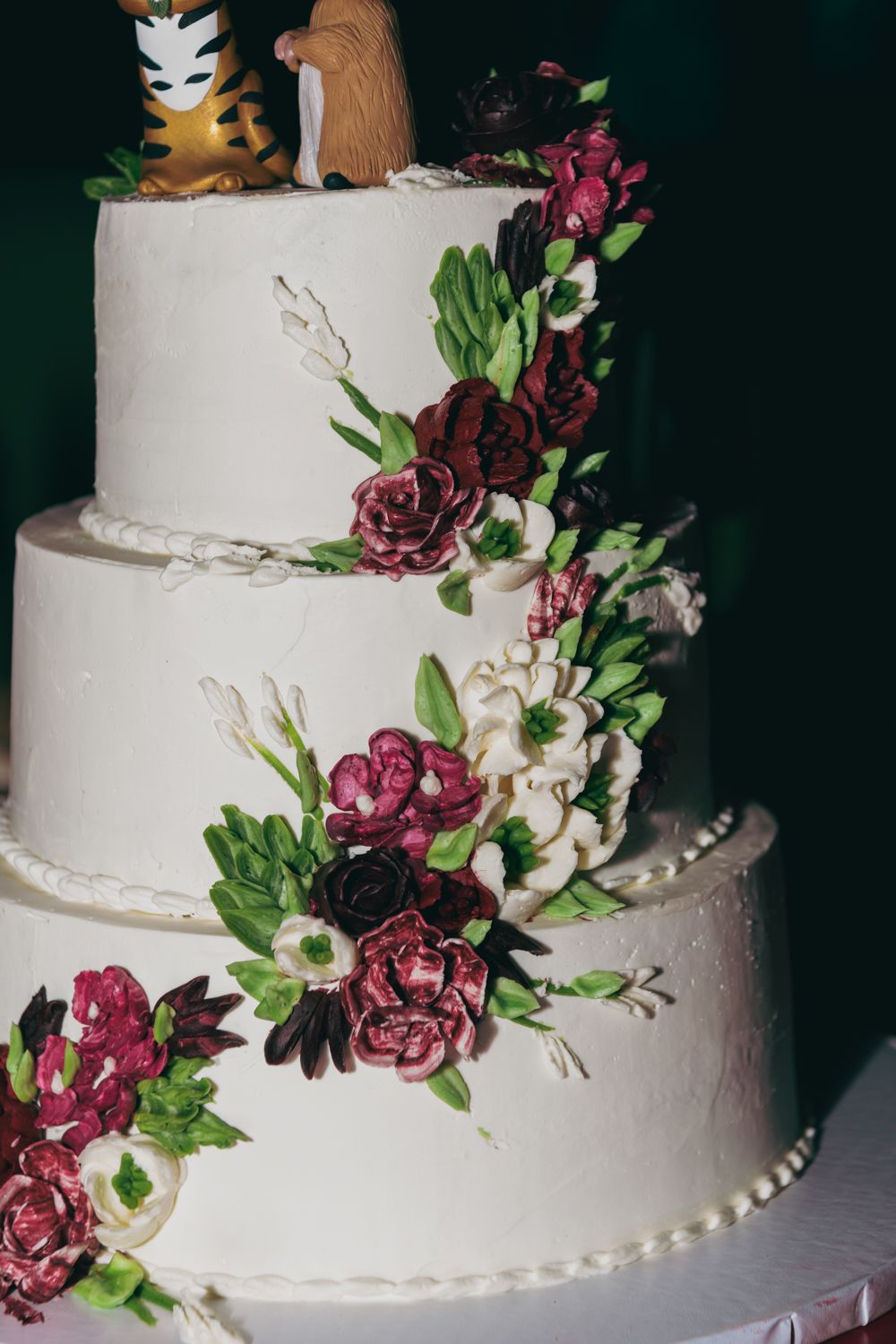Three-tier white wedding cake adorned with burgundy and white sugar flowers and green leaves.