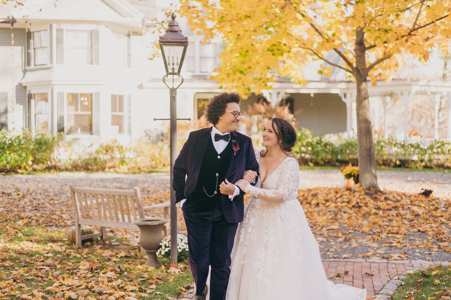 Bride and groom walking hand in hand among golden autumn leaves outside a white house.