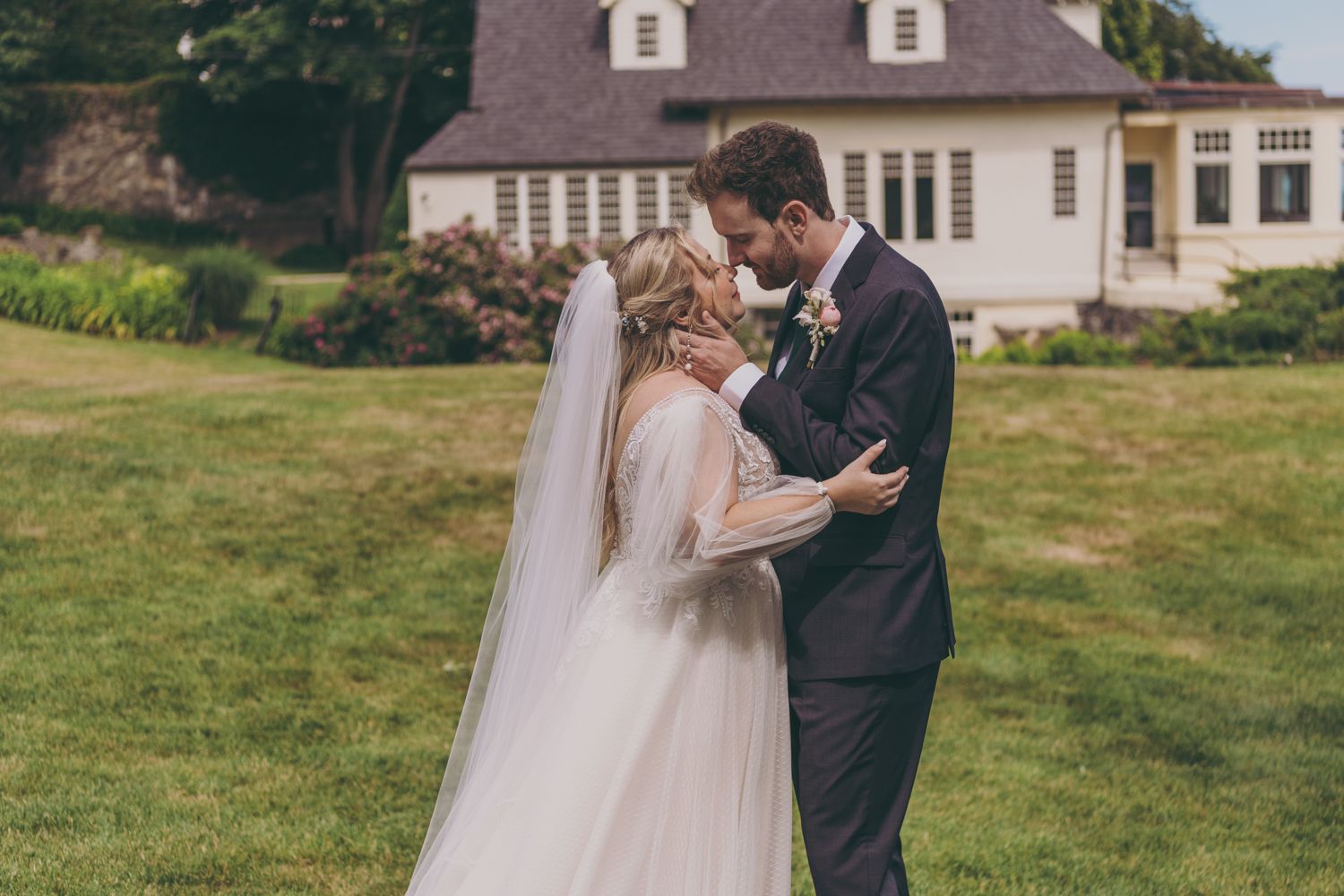 A bride and groom share a tender moment embracing near a white manor house on their wedding day.