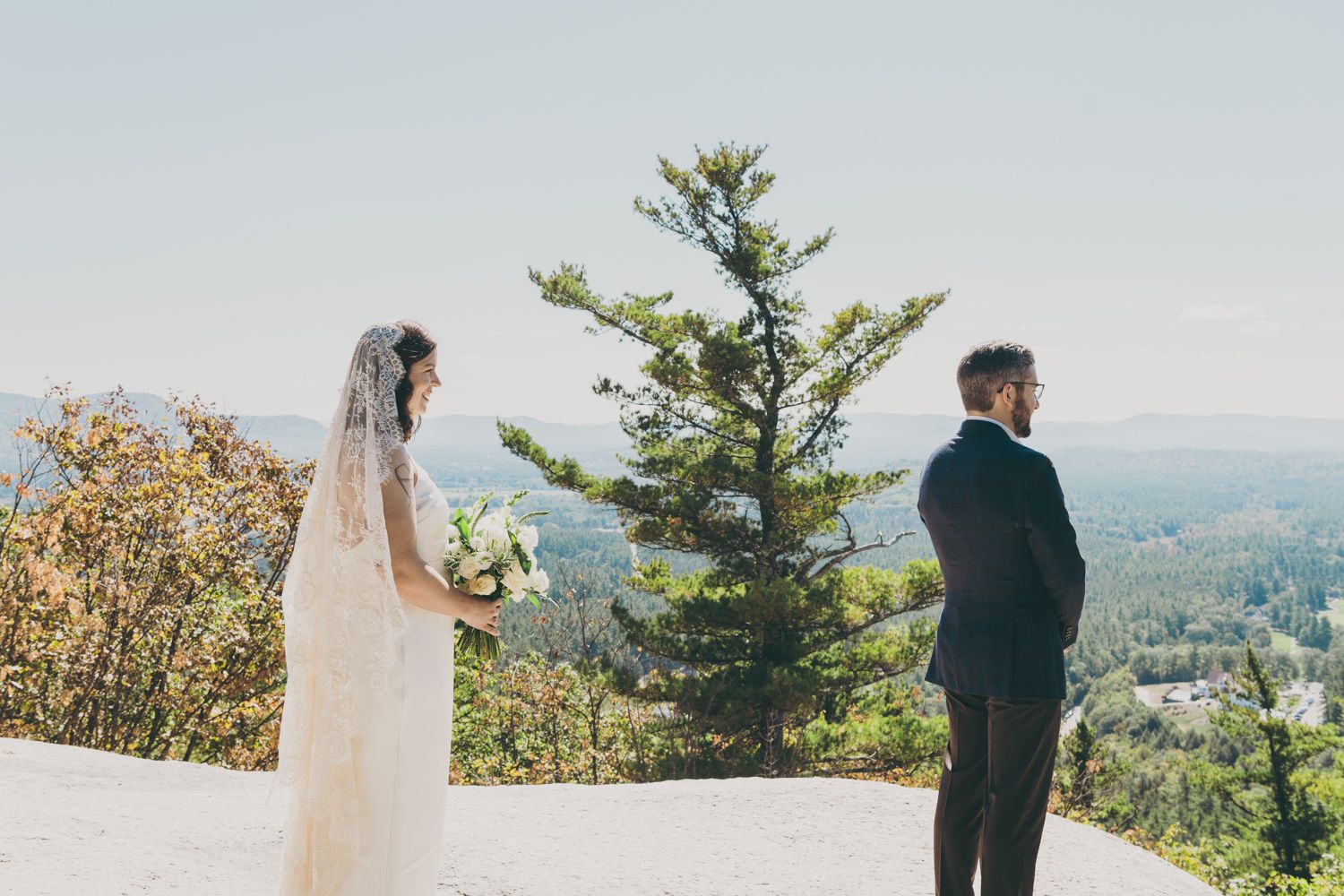 Bride in white gown with veil and groom standing apart on rocky mountain summit first look.