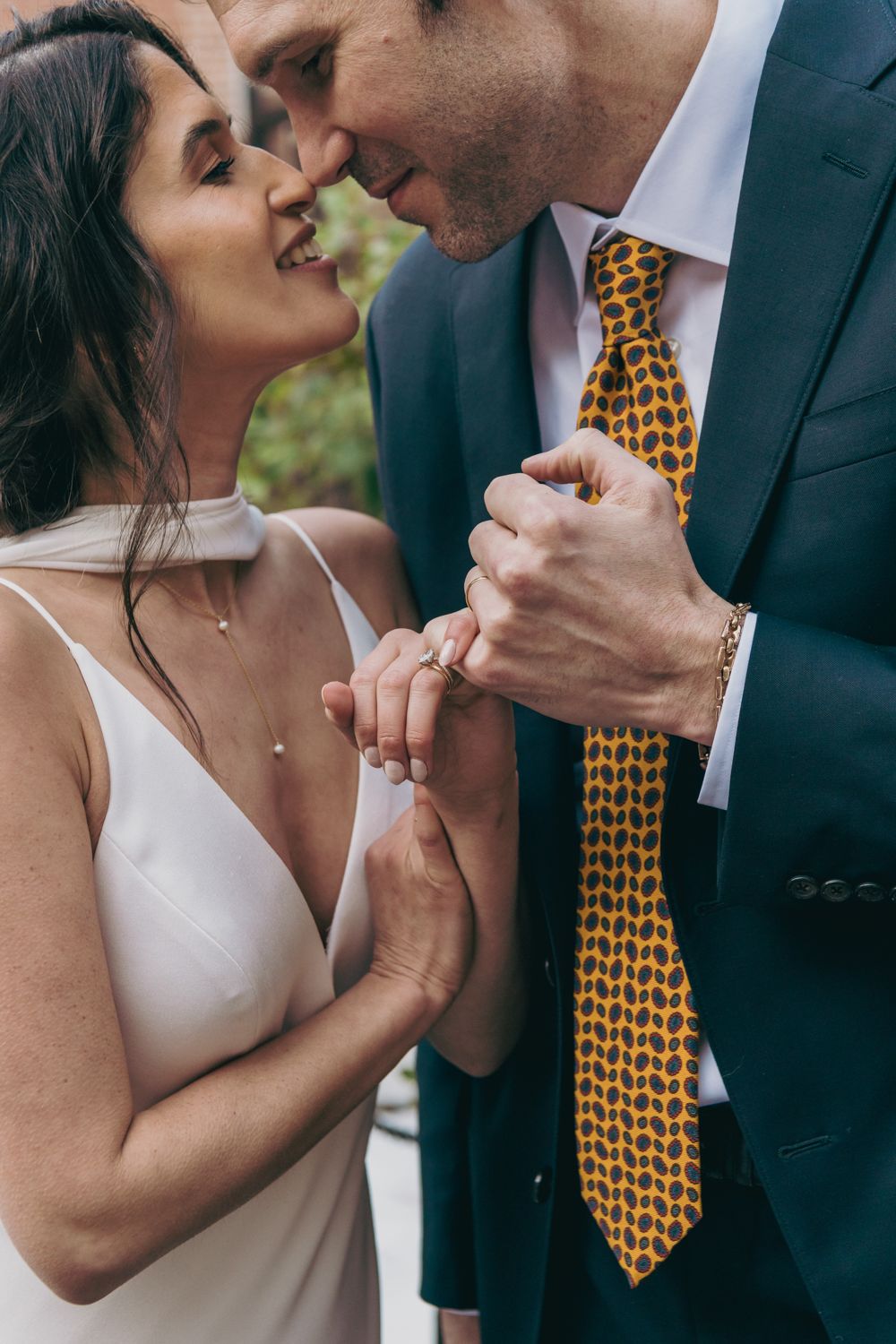 Bride and groom nearly kissing, she in white dress, he in navy suit with yellow patterned tie, holding hands.