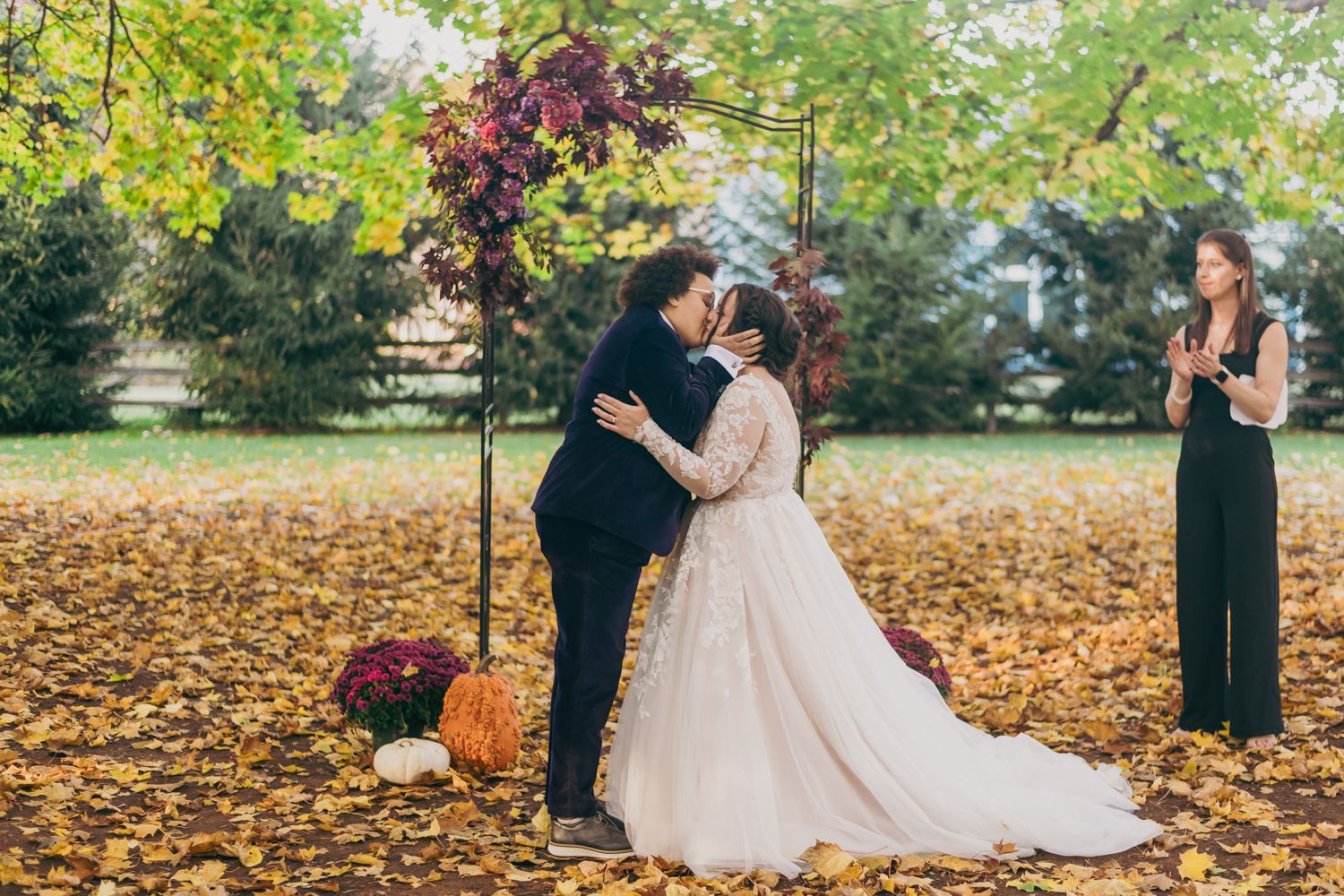 Newlyweds share first kiss under floral arch surrounded by golden autumn leaves.